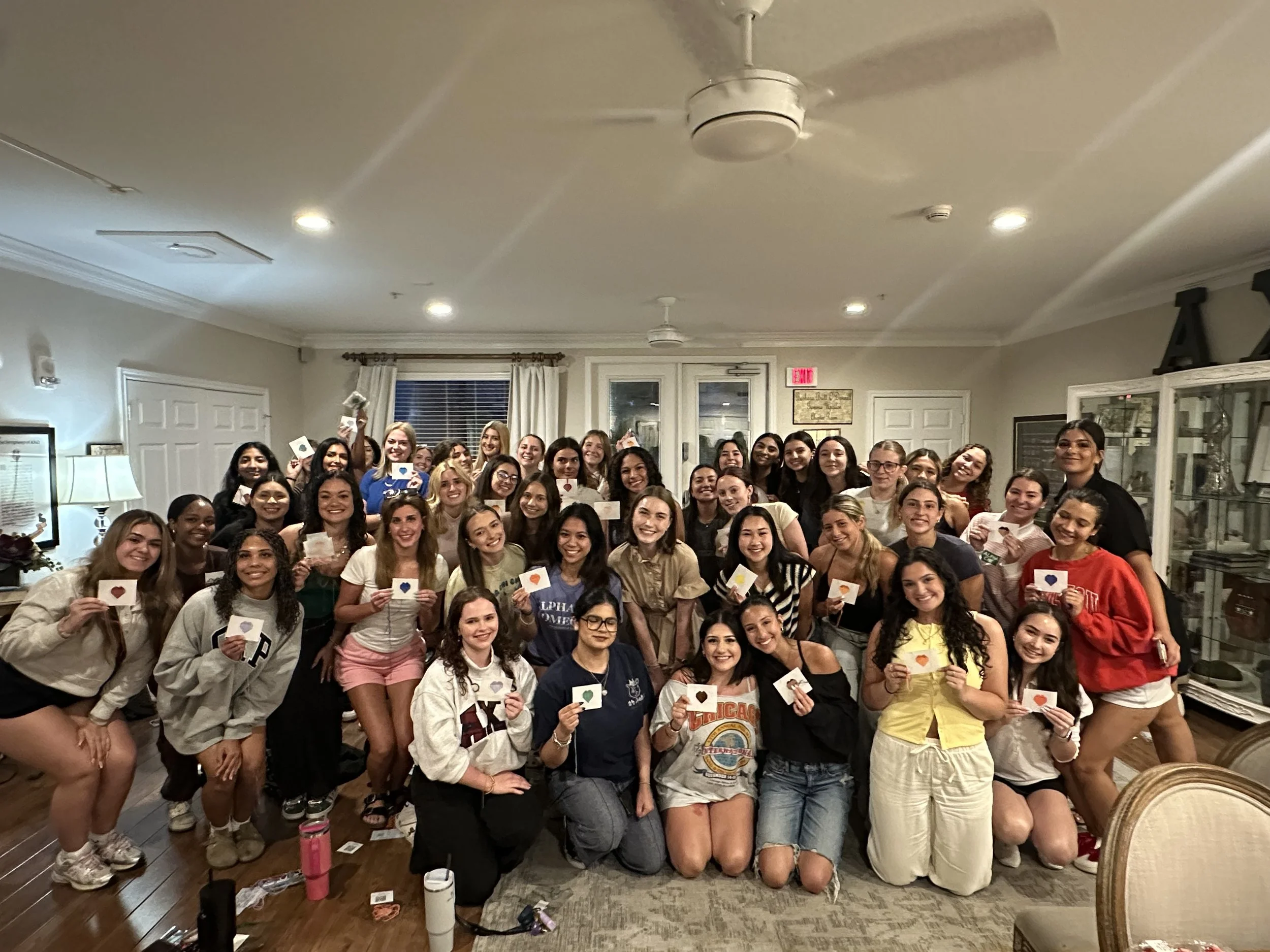 A large group of smiling young women and a few men poses together indoors, holding small cards with colored hearts on them, in a warmly lit room with wooden floors, curtains, and furniture.