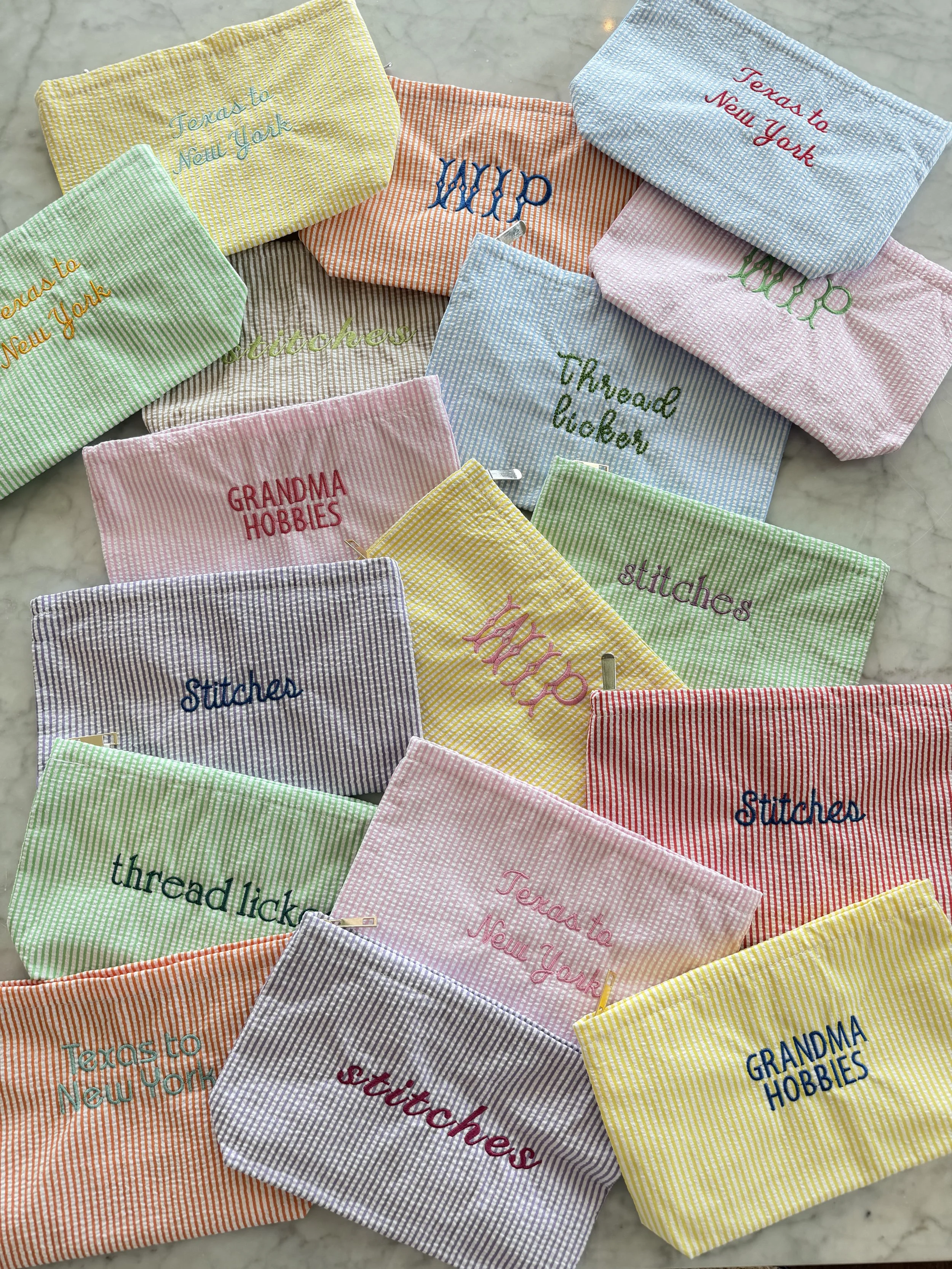 Multiple colorful embroidered zippered pouches with various phrases like "Tens to New York," "Grandma Hobbies," "Stacks," and "Threaded Lucky" on a marble surface.