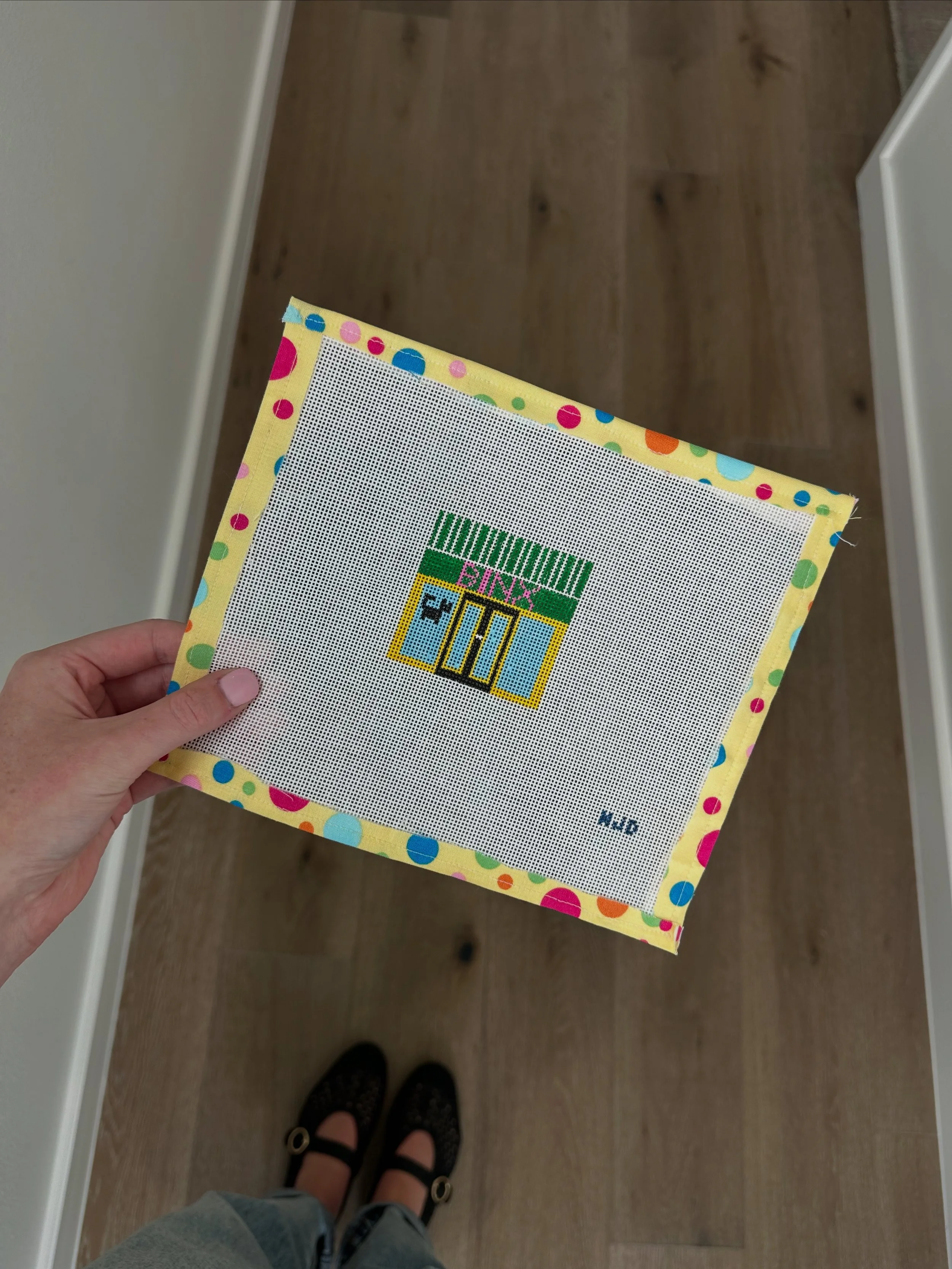 Person holding a colorful cross-stitch pattern in front of them, with their feet in black shoes visible on a wooden floor.