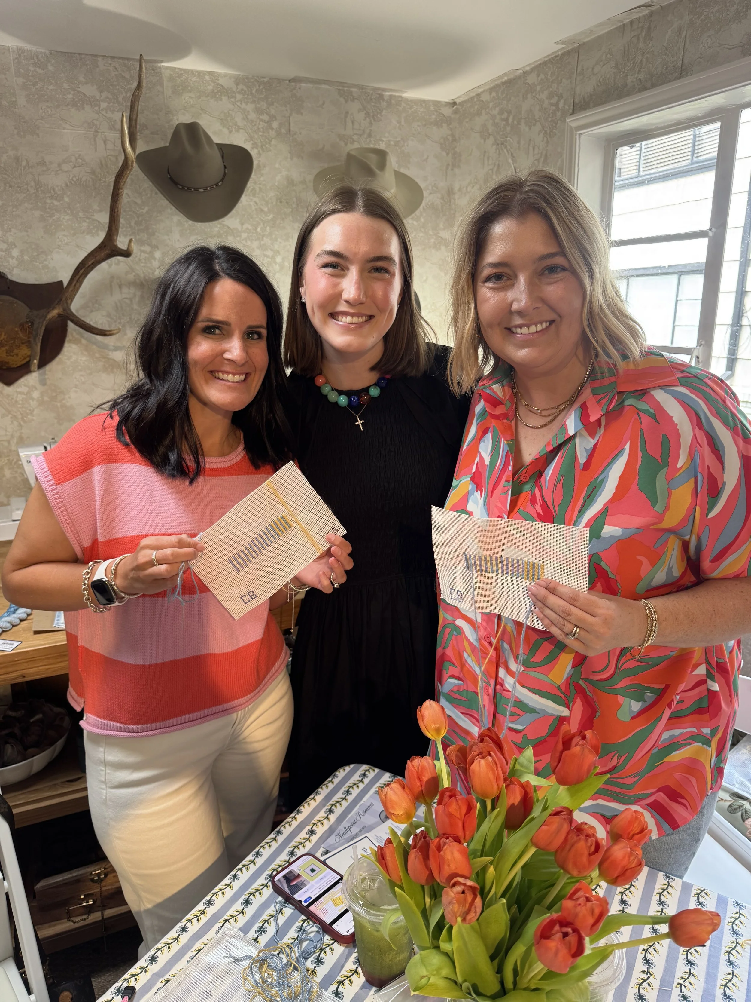 Three women standing indoors, smiling at the camera, holding small fabric swatches, with a table of flowers, snacks, and a smartphone in front of them.