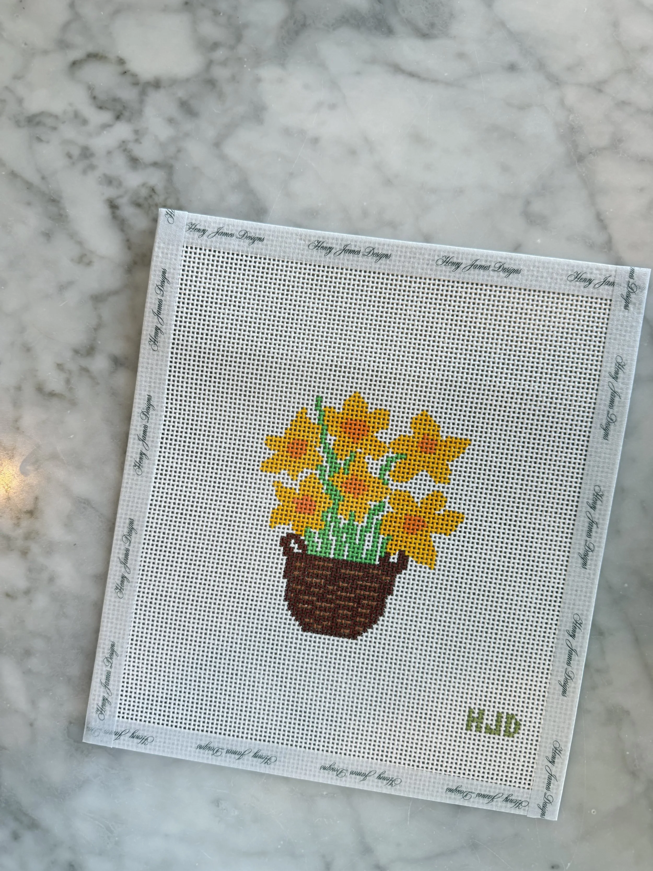 Cross-stitch embroidery of a potted yellow flower with green leaves on a white grid fabric.