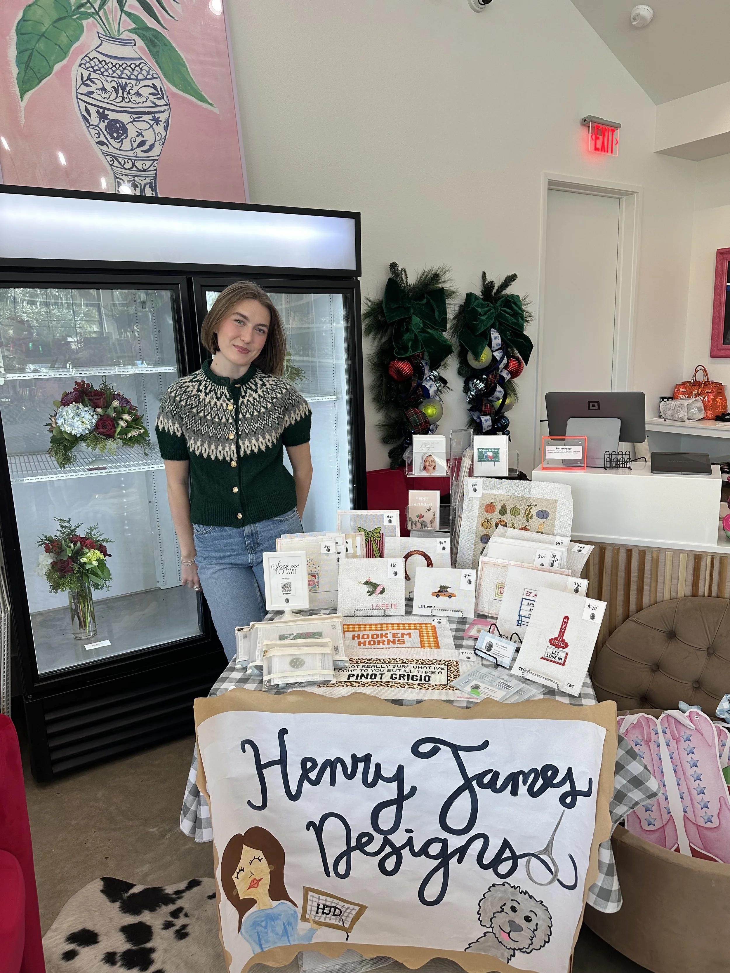 A woman with shoulder-length brown hair, wearing a green and white patterned sweater, stands in front of a refrigerated display case filled with bouquets of flowers. Decorative Christmas wreaths hang on the wall, and a table in the foreground display