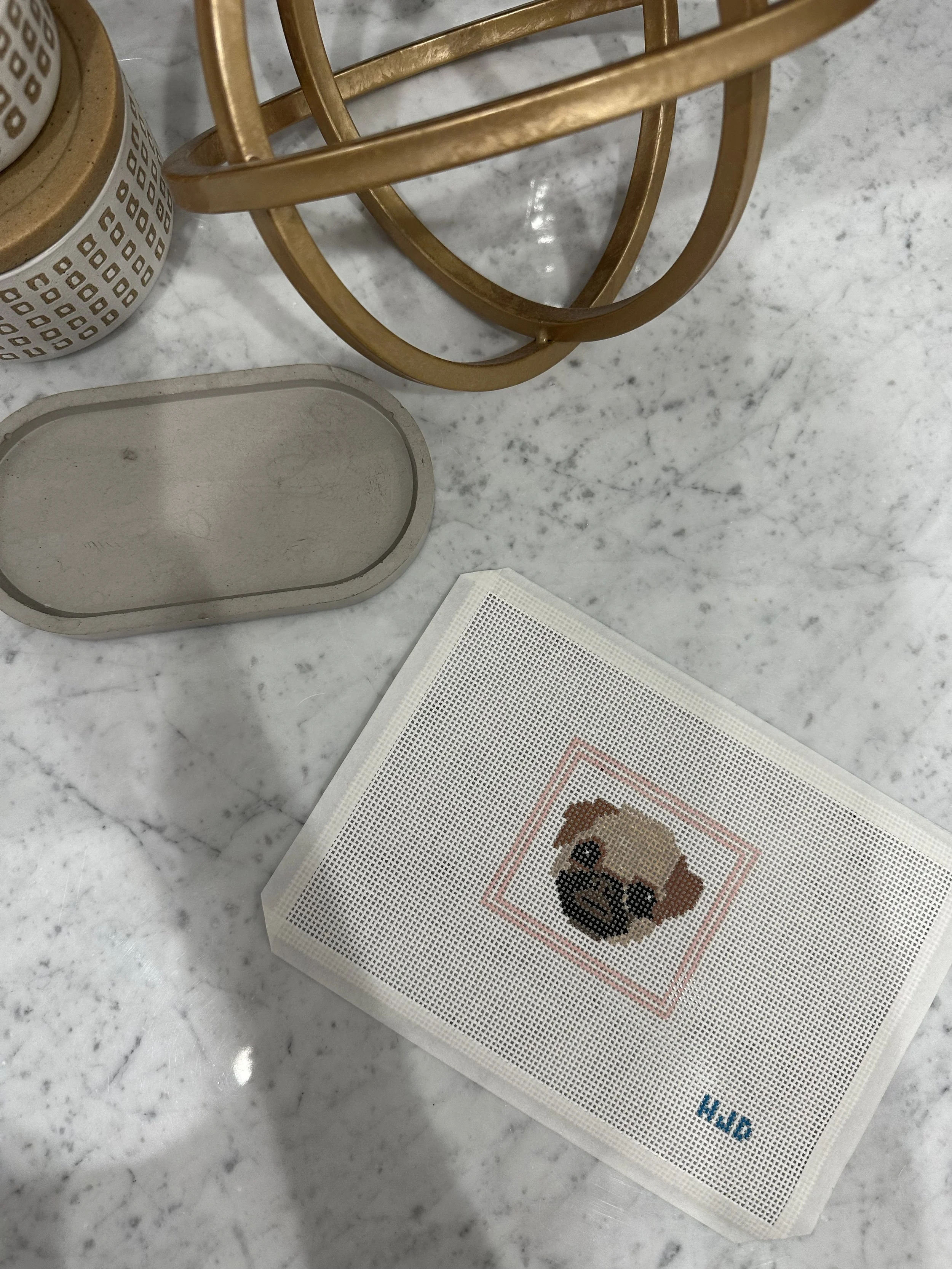 A close-up of a marble countertop with a gold metal decorative object, a white rectangular object with a pixelated dog image, a ceramic container with a lattice pattern, and a mesh bag with the word 'KIND' printed on it.