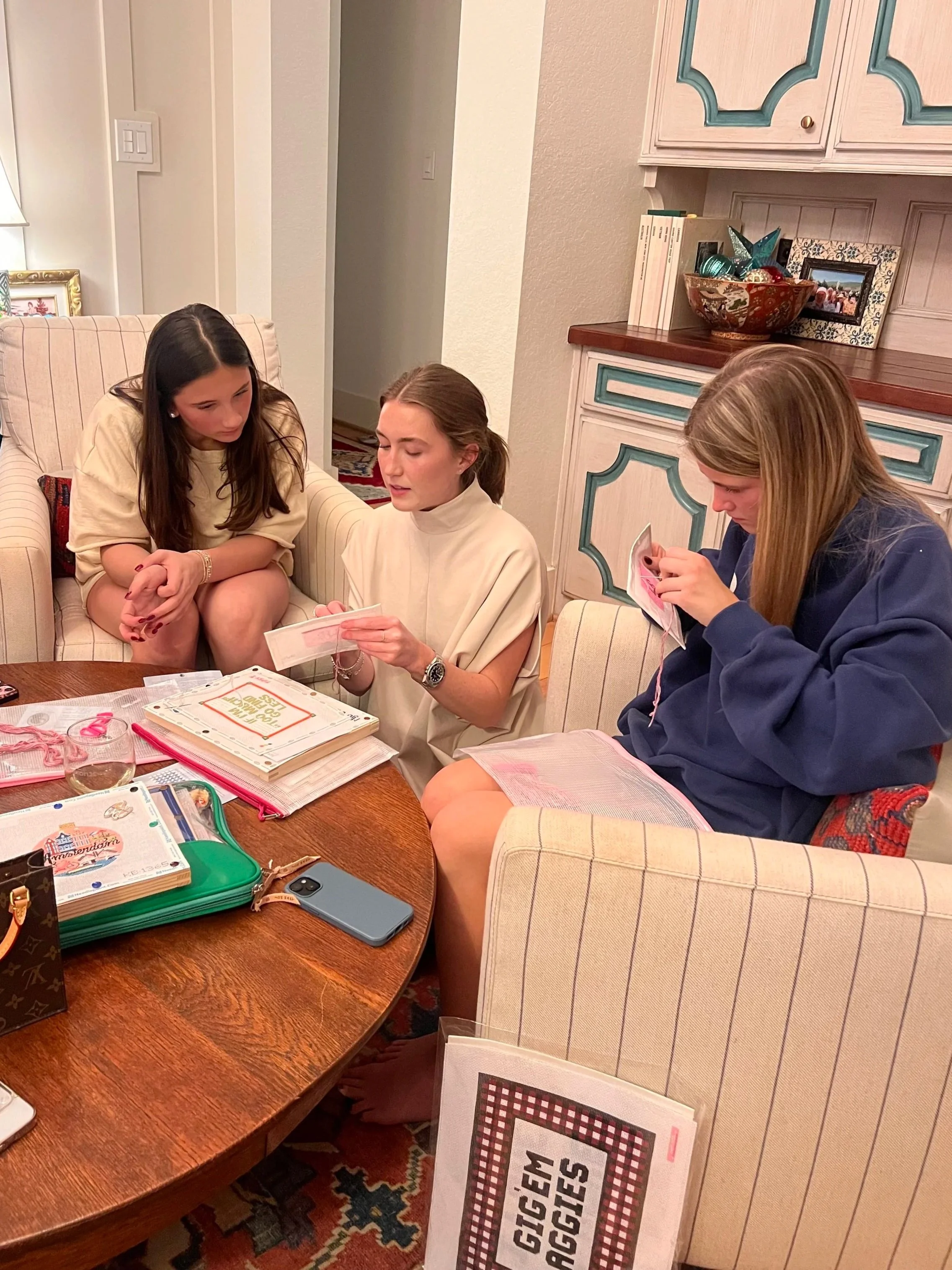 Three women sitting around a wooden table, looking at papers and a book, in a cozy room. One woman is reading a paper, the second is looking at a booklet, and the third is examining a piece of paper. Various books, a phone, and personal items are on the table.