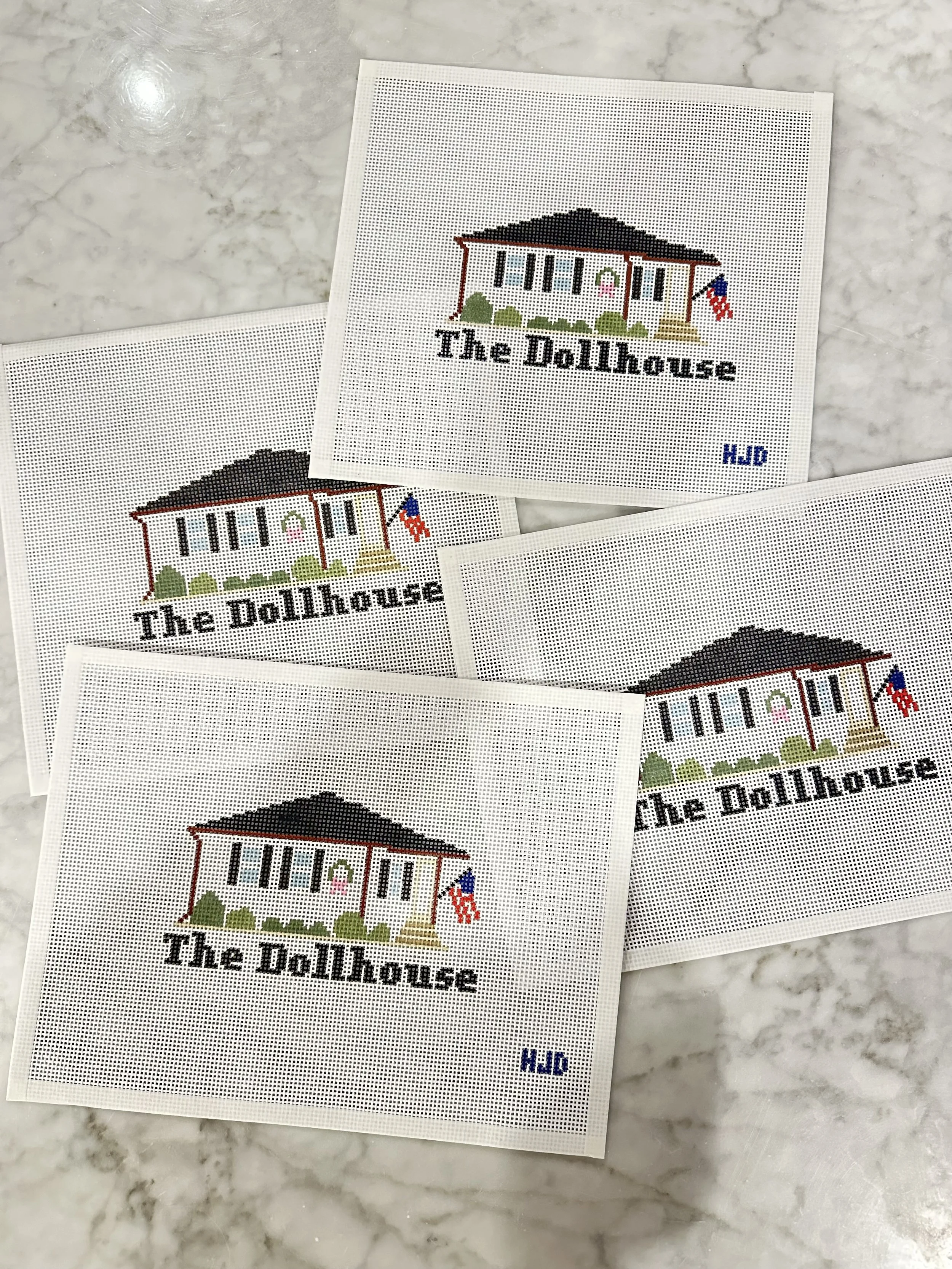 Multiple needlepoint cross-stitch pieces featuring a dollhouse with a porch, windows, a door, and a flag, laid on a marble surface.
