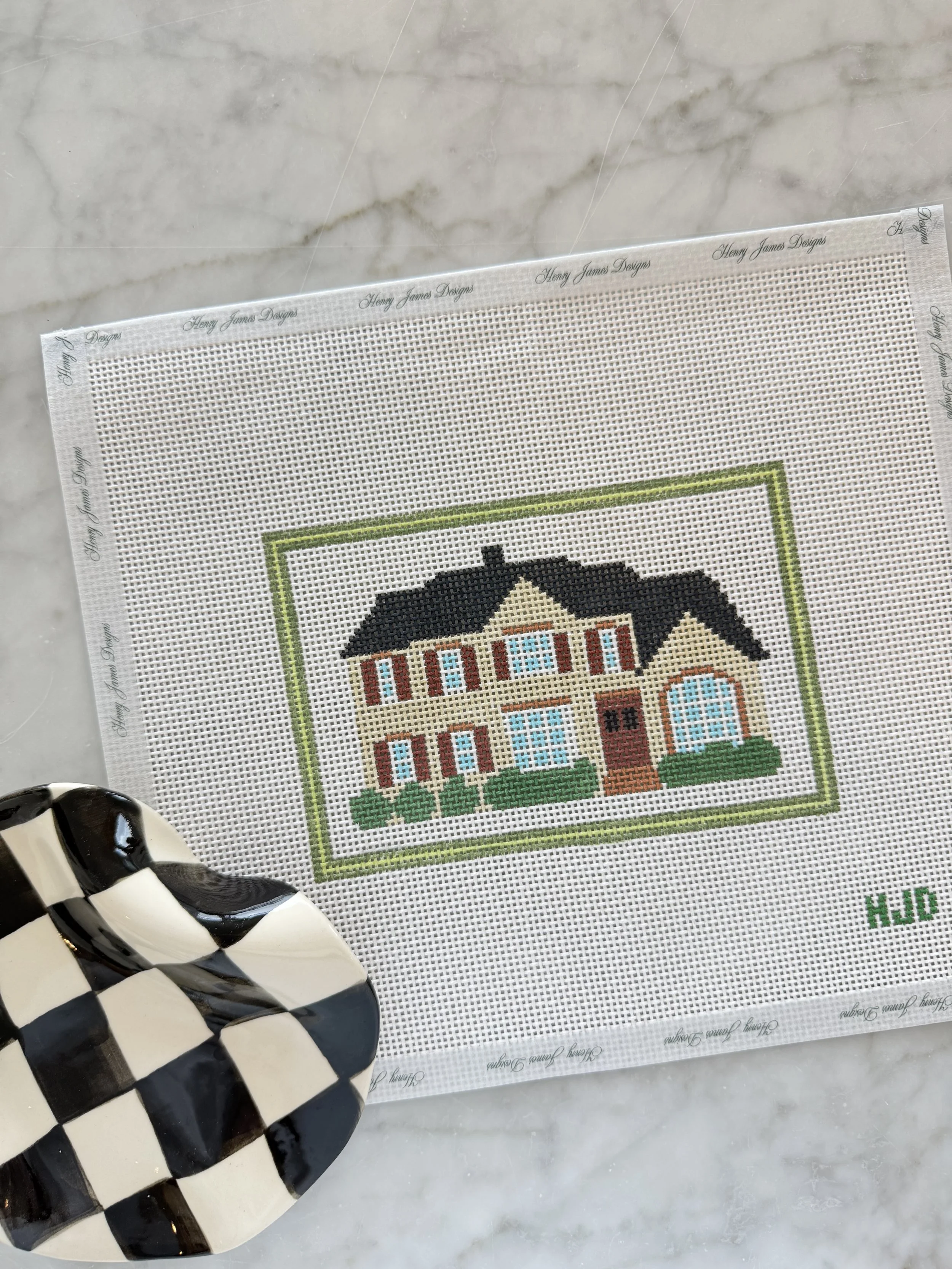 Part of a cross-stitched picture of a house with a green lawn, brick chimney, multiple windows, and a black roof on embroidery canvas. A black and white checkered ceramic vase is in the foreground.
