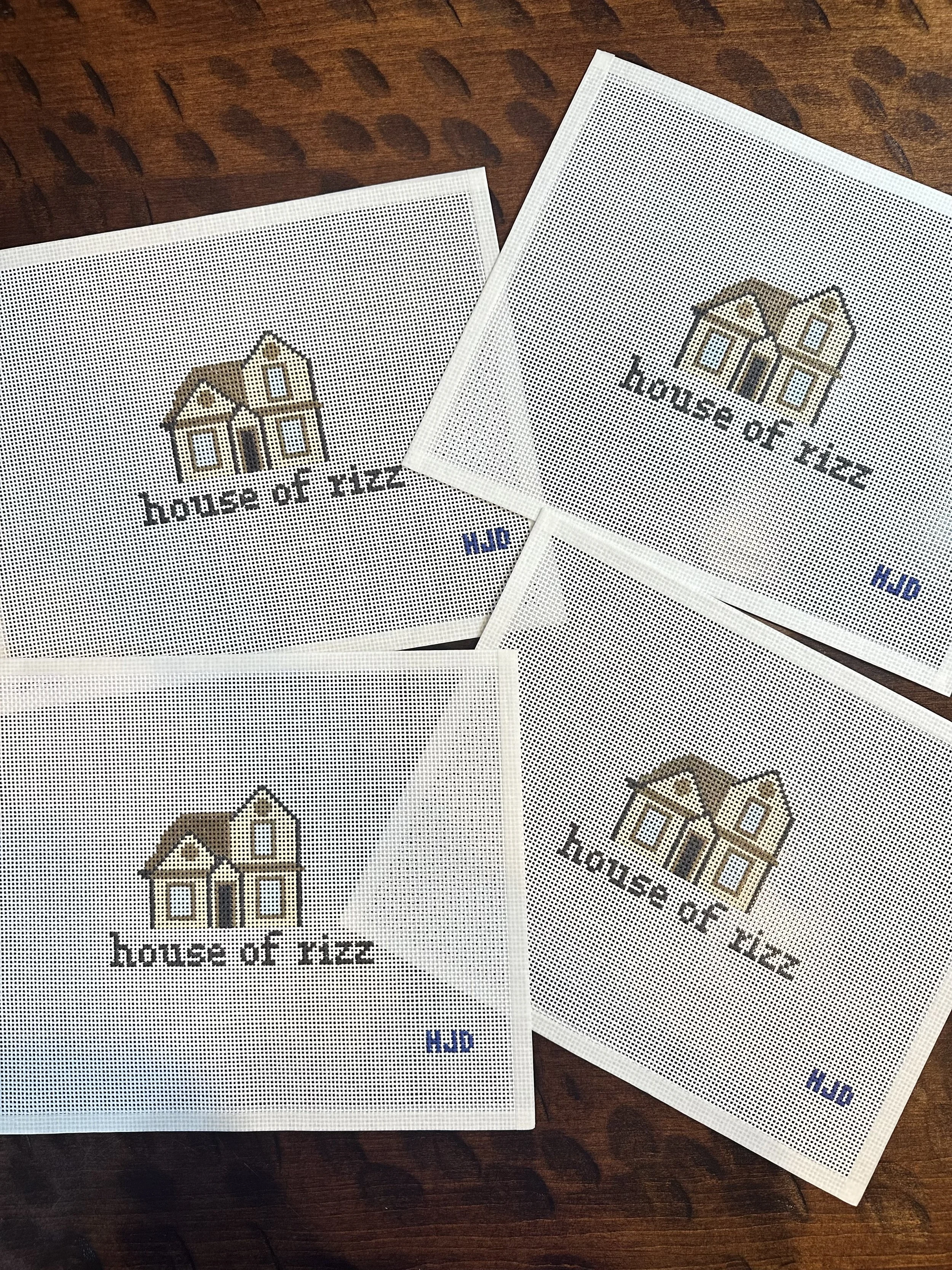 Two perforated cross-stitch patterns with a house design and the text 'house of tiles' and 'HOD' on a dark wooden surface.