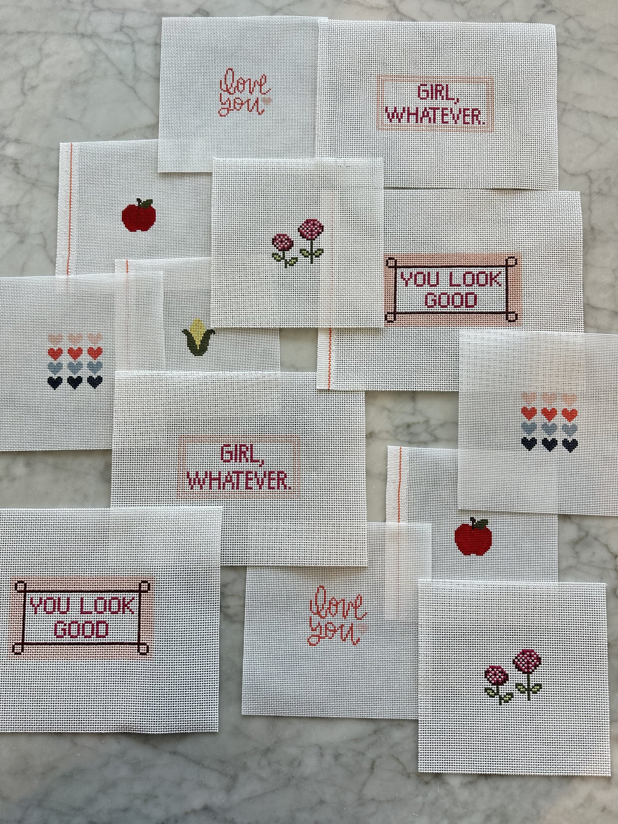 Assorted cross-stitch embroidery pieces with various designs including hearts, flowers, a red apple, and phrases like 'GIRL, WHATEVER,' 'YOU LOOK GOOD,' and 'love you.'