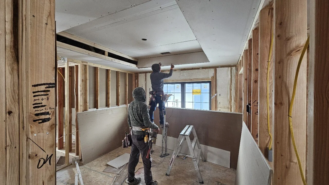 Construction workers installing drywall in an unfinished room with exposed wooden framing and electrical wires.
