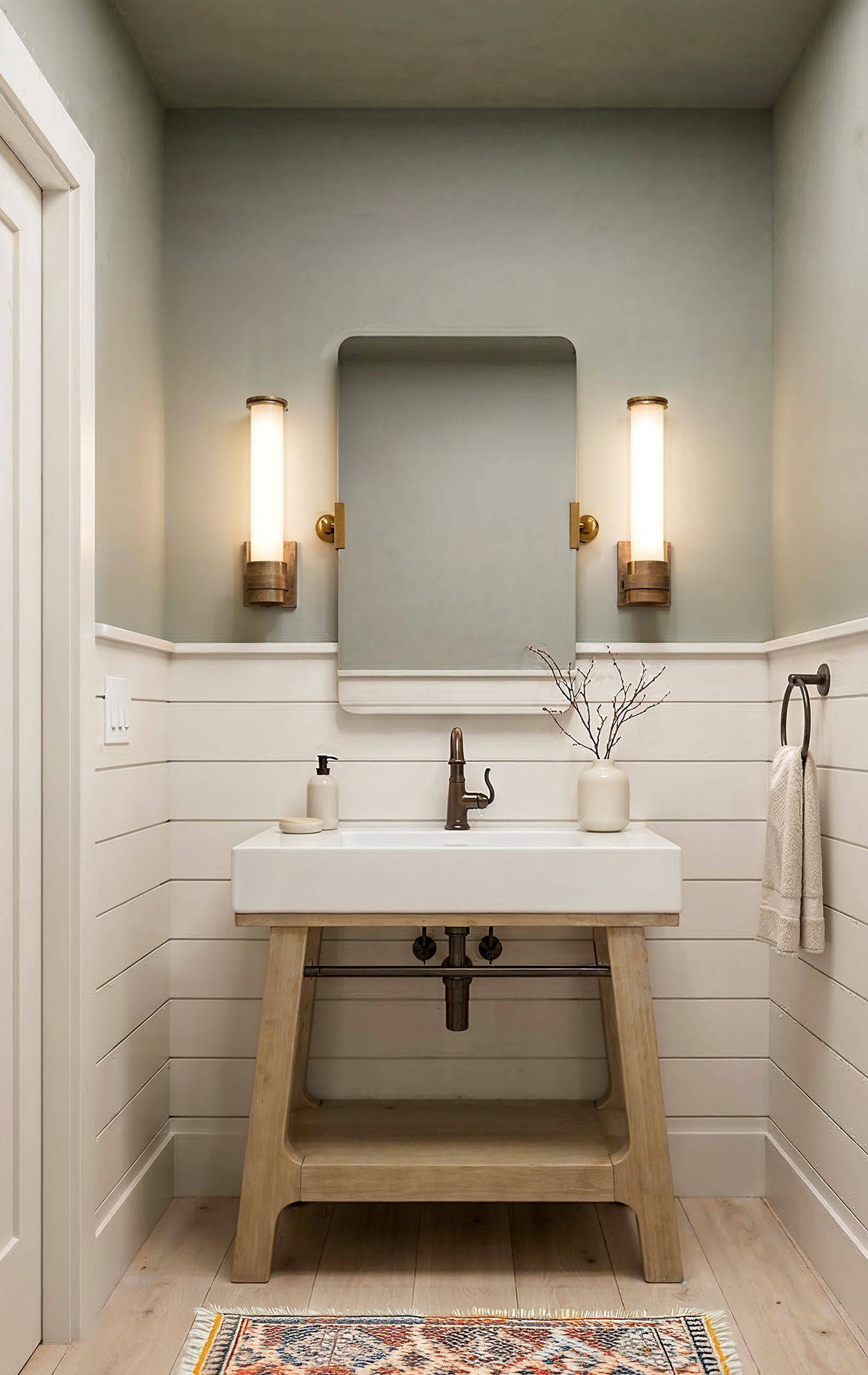 A powder room in a Sloan home with sage green walls, white shiplap wainscoting, brass tube wall sconces, and a natural wood vanity with white trough sink