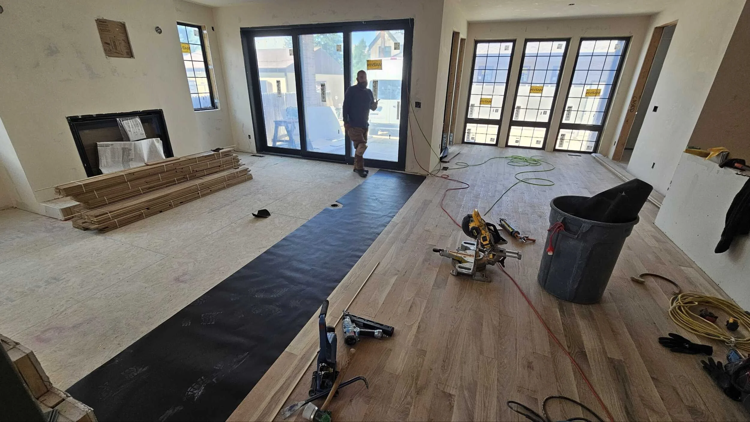 Interior of a house under construction with flooring being installed. A worker is by a large sliding glass door, with construction materials, tools, and wiring scattered around.