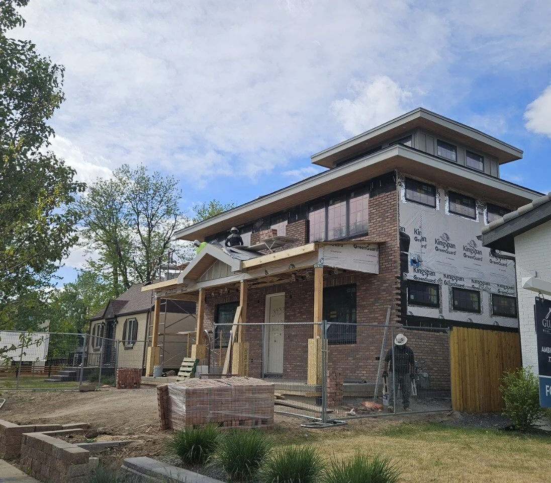 A two-story house under construction with workers installing the roof. The house has a brick exterior, and scaffolding surrounds parts of the structure.