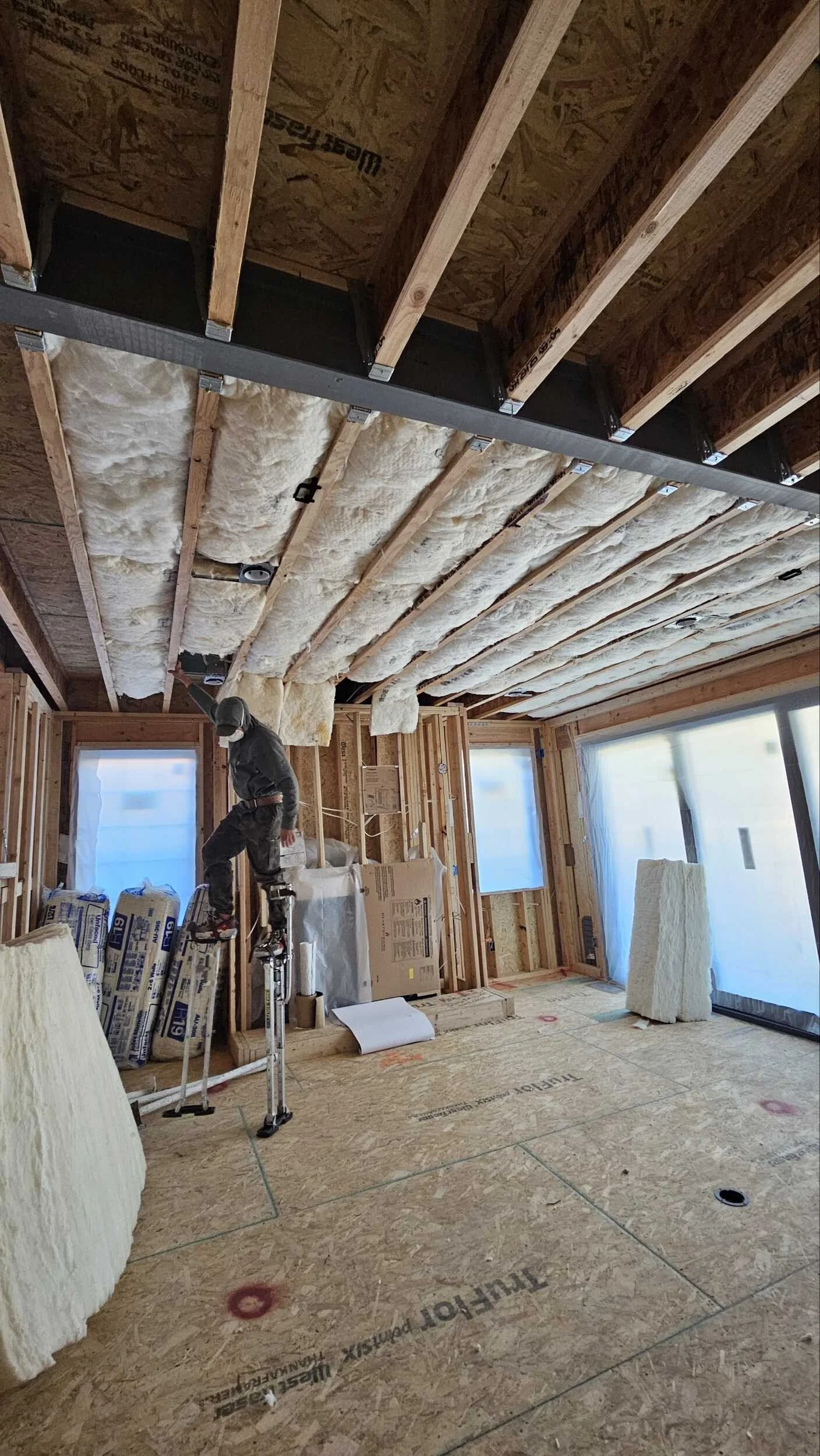 Interior of a house under construction, showing exposed wooden framing, insulation, and windows. A worker is standing on a small ladder, working on the ceiling. Building materials and tools are scattered across the floor.