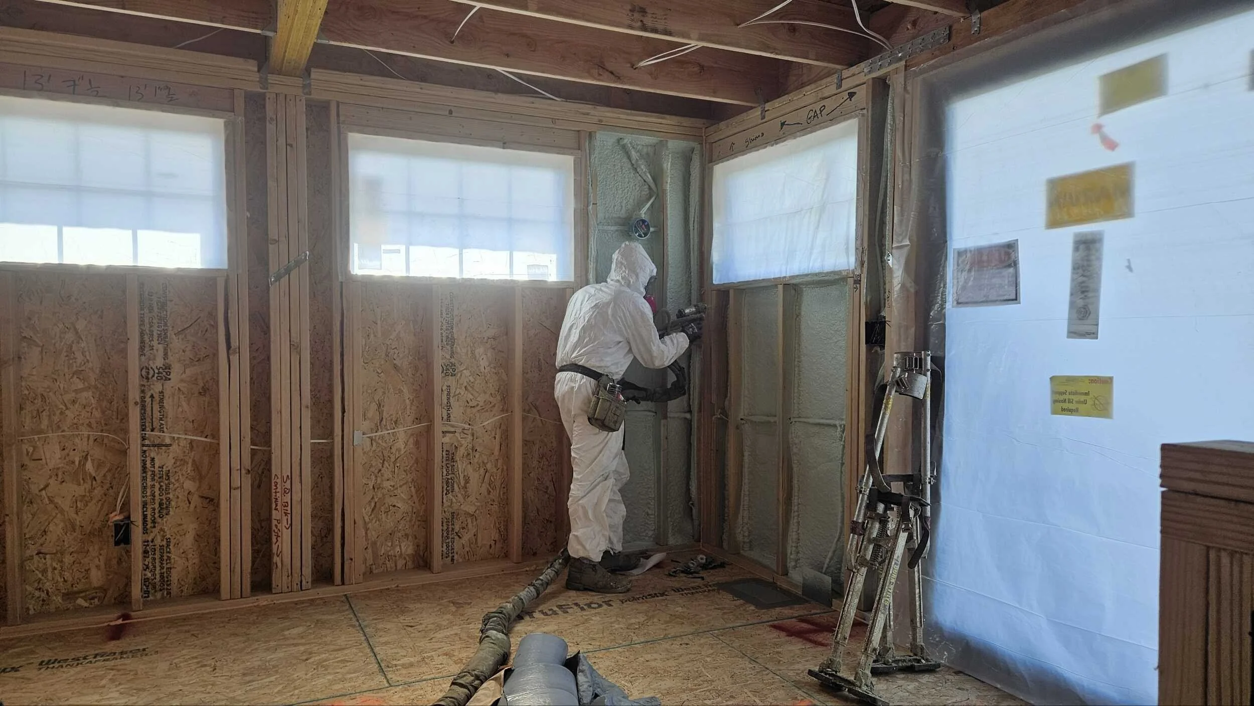 Worker in white protective suit and mask spray-foaming a wall inside a construction site. The wall has three windows covered with plastic, and there is construction equipment on the floor, including a ladder and hoses.
