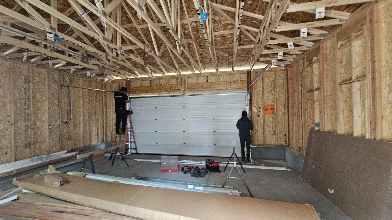 Interior of a garage under construction with two workers installing a garage door. One worker is on a ladder, and the other is standing on the ground. Building materials and tools are scattered around.