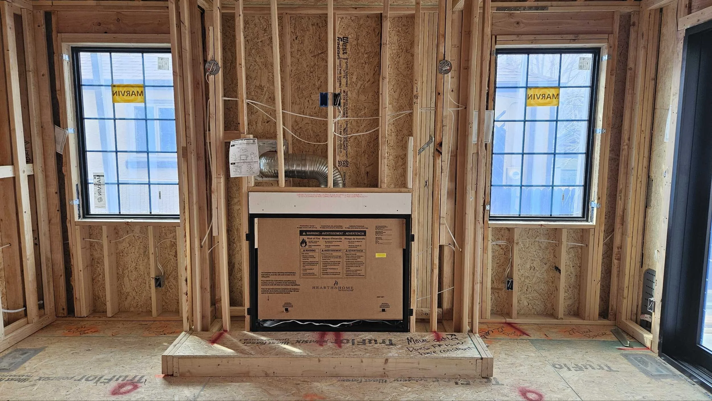 Interior view of a room under construction with exposed wooden framing, two large windows, and an HVAC unit in the center.