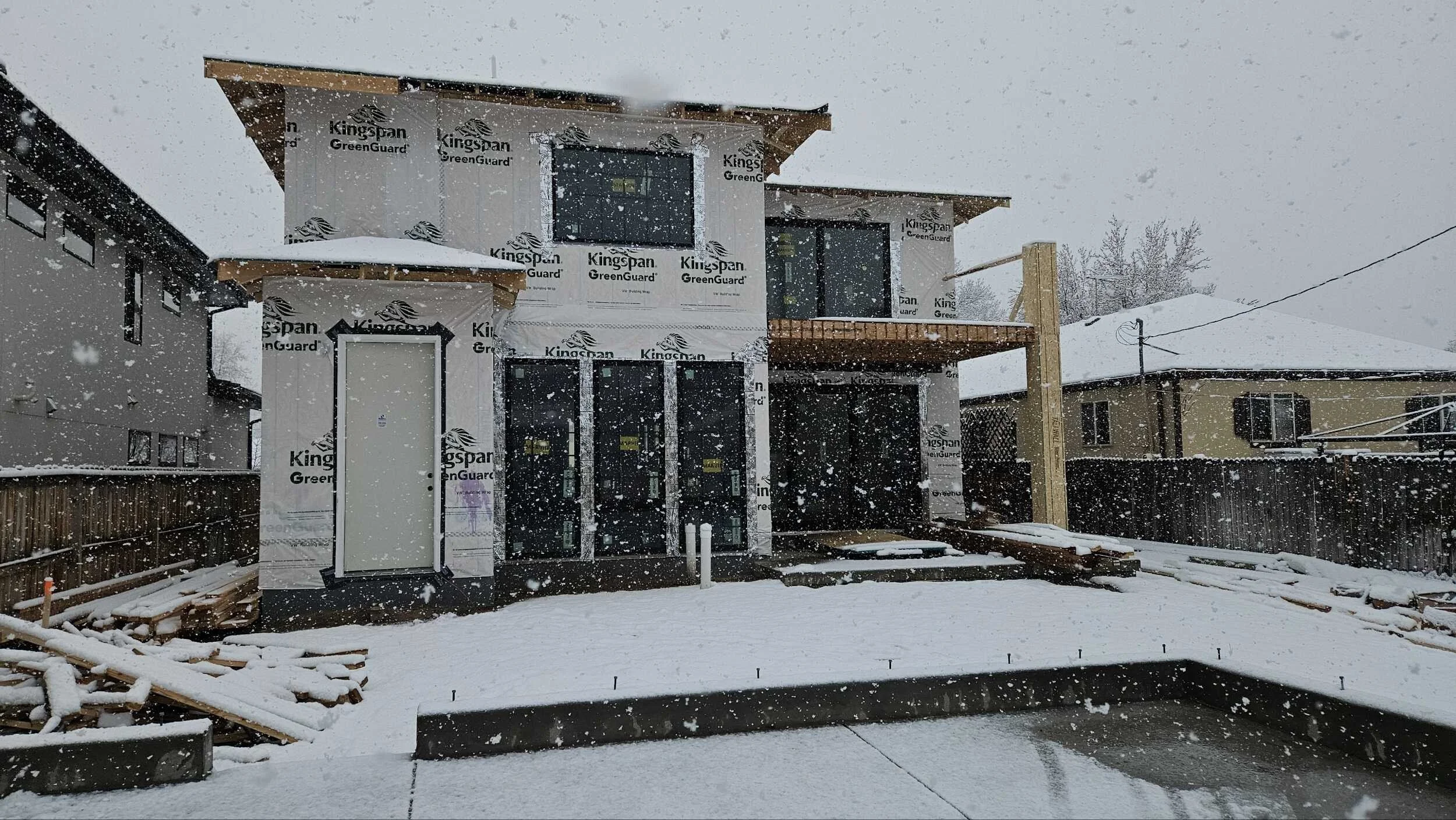 Newly constructed house under snow with protective wrapping on exterior walls, a deck, and a backyard fenced area.