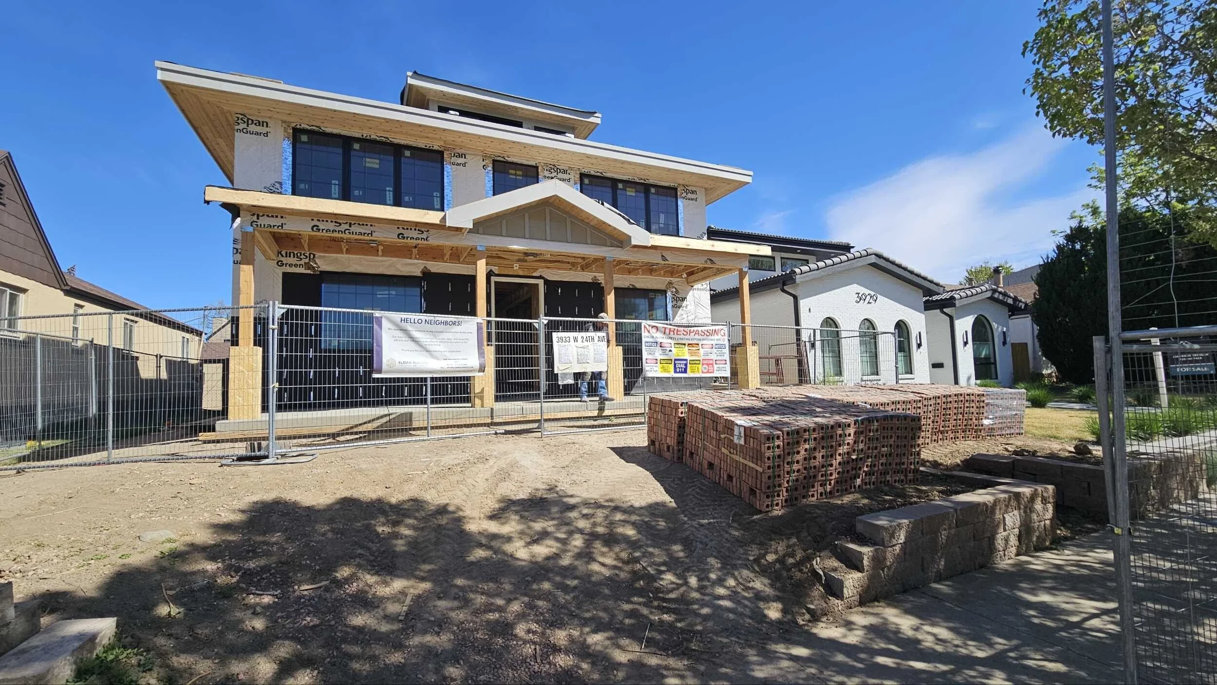 A house under construction with a wooden frame and black windows, surrounded by a fence with a sign, with neighboring houses visible and a clear blue sky above.