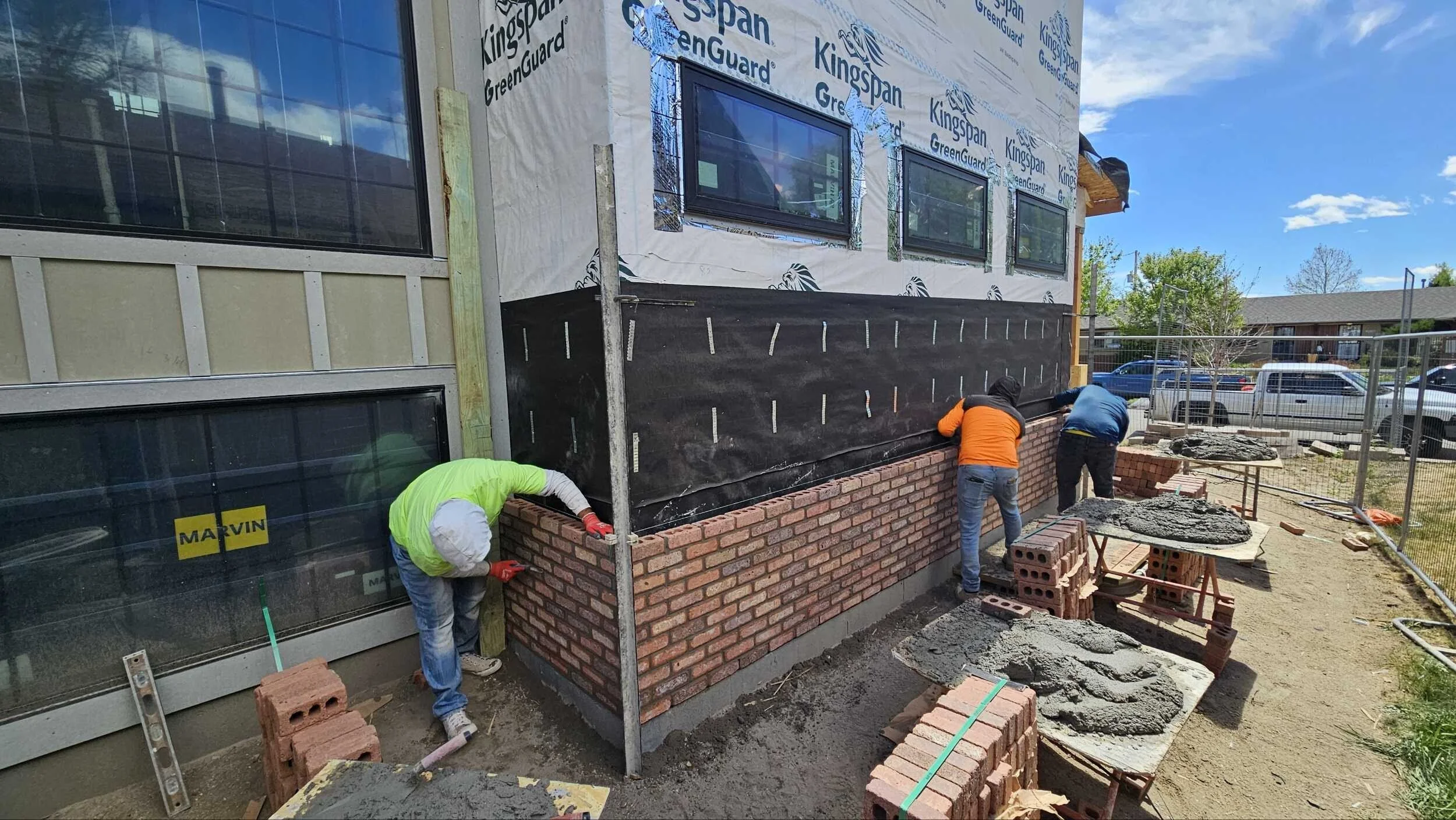 Construction workers building a brick wall outside a house with window installation, under a bright blue sky.
