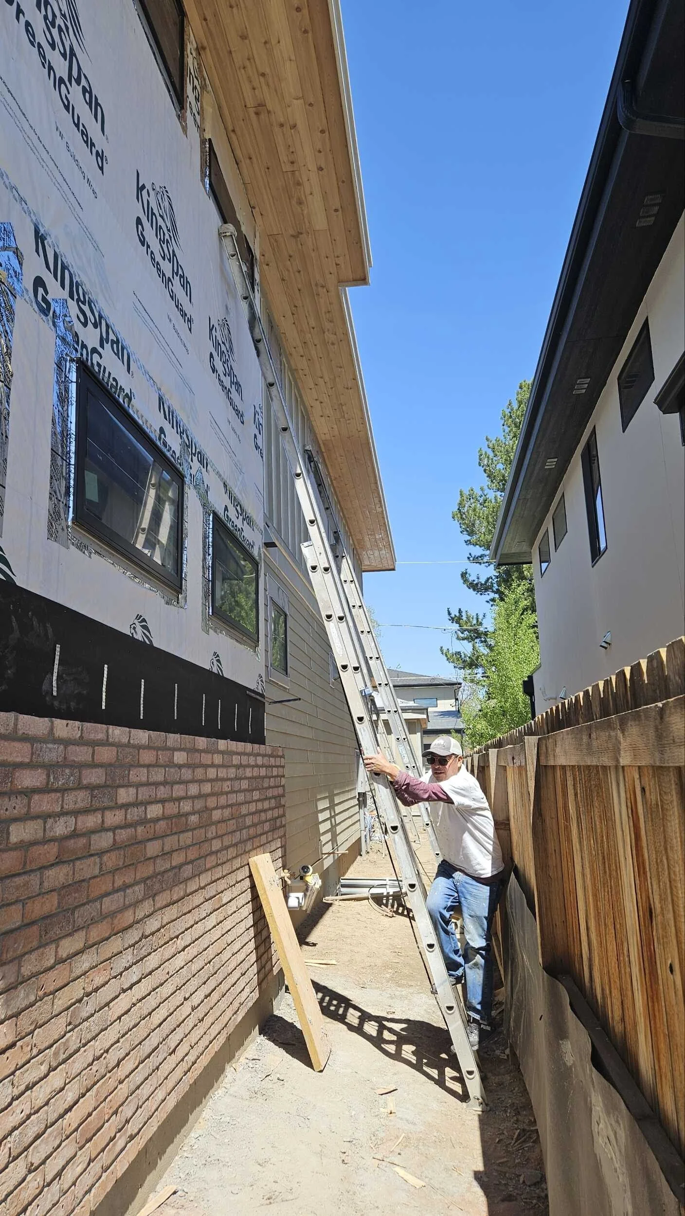 A man standing on a ladder working on the exterior of a house under construction, with siding and windows, during daytime, with a clear blue sky and a wooden fence.