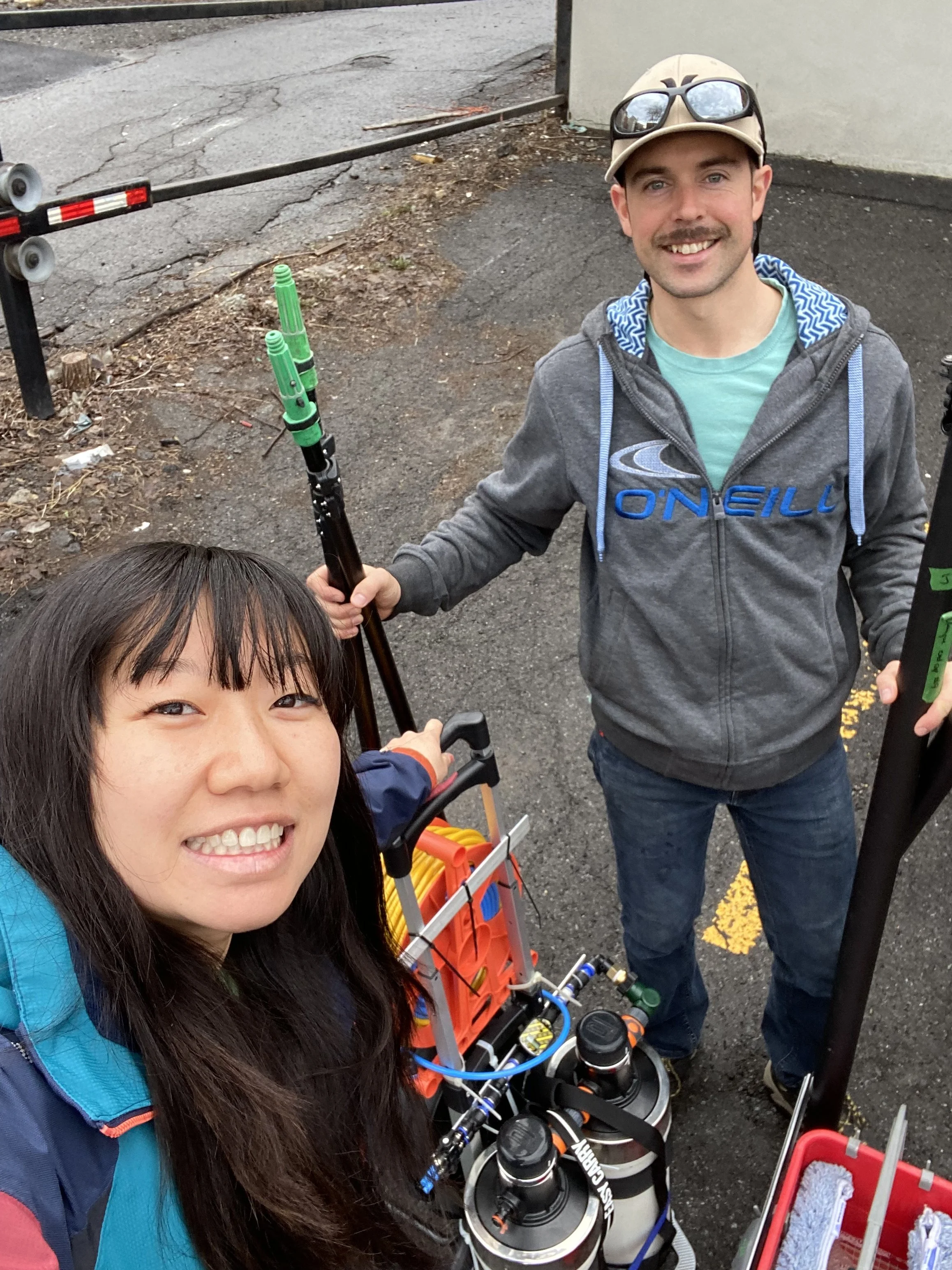 Two people smiling for a selfie on a street, with equipment related to scuba diving, including tanks, a crate with snorkeling gear, and a tow cart, possibly preparing for a diving activity.