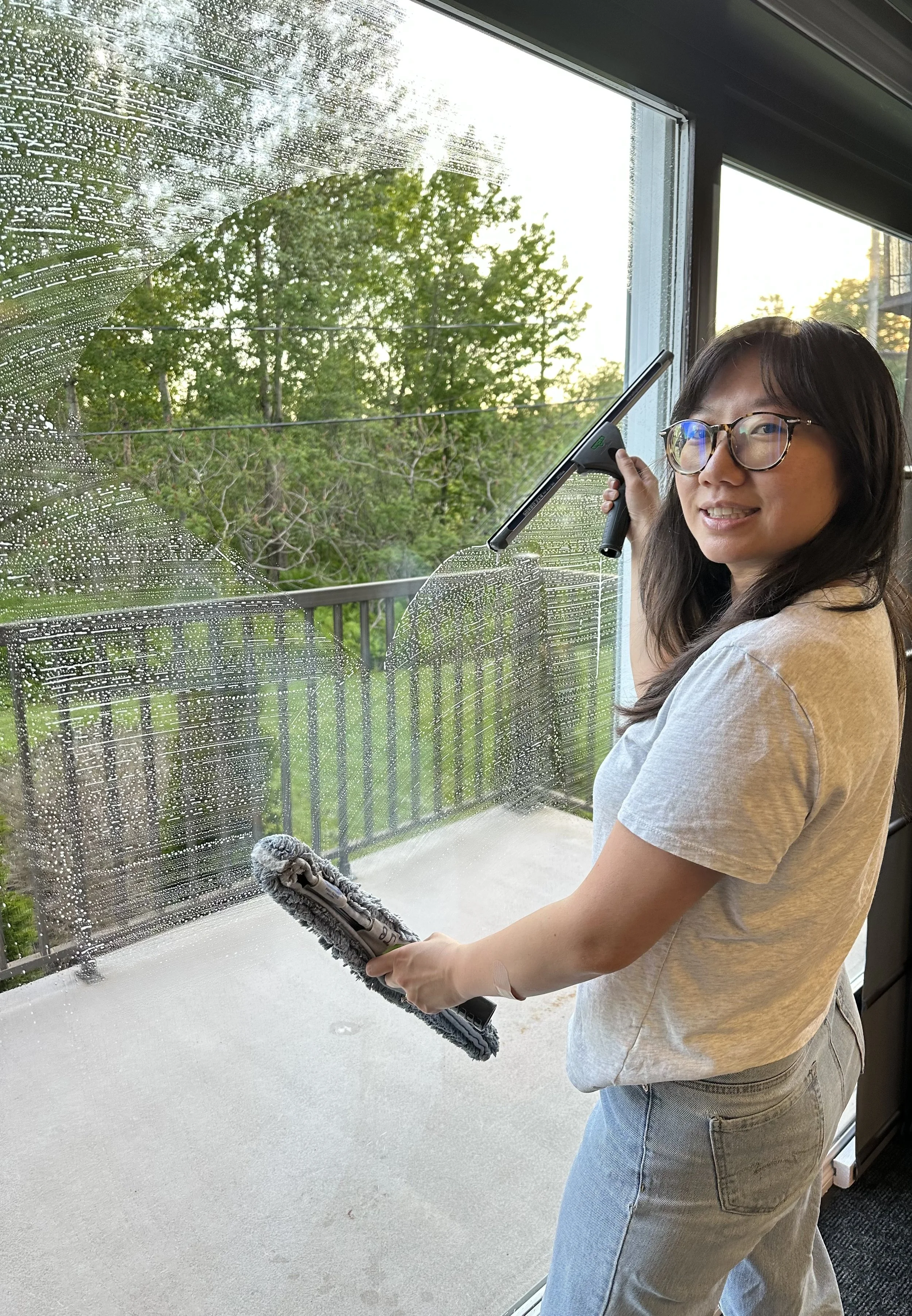 Woman cleaning a window with a squeegee, wearing glasses and a beige t-shirt, standing inside a house during daylight. The window shows rain droplets and a green outdoor scenery with trees.