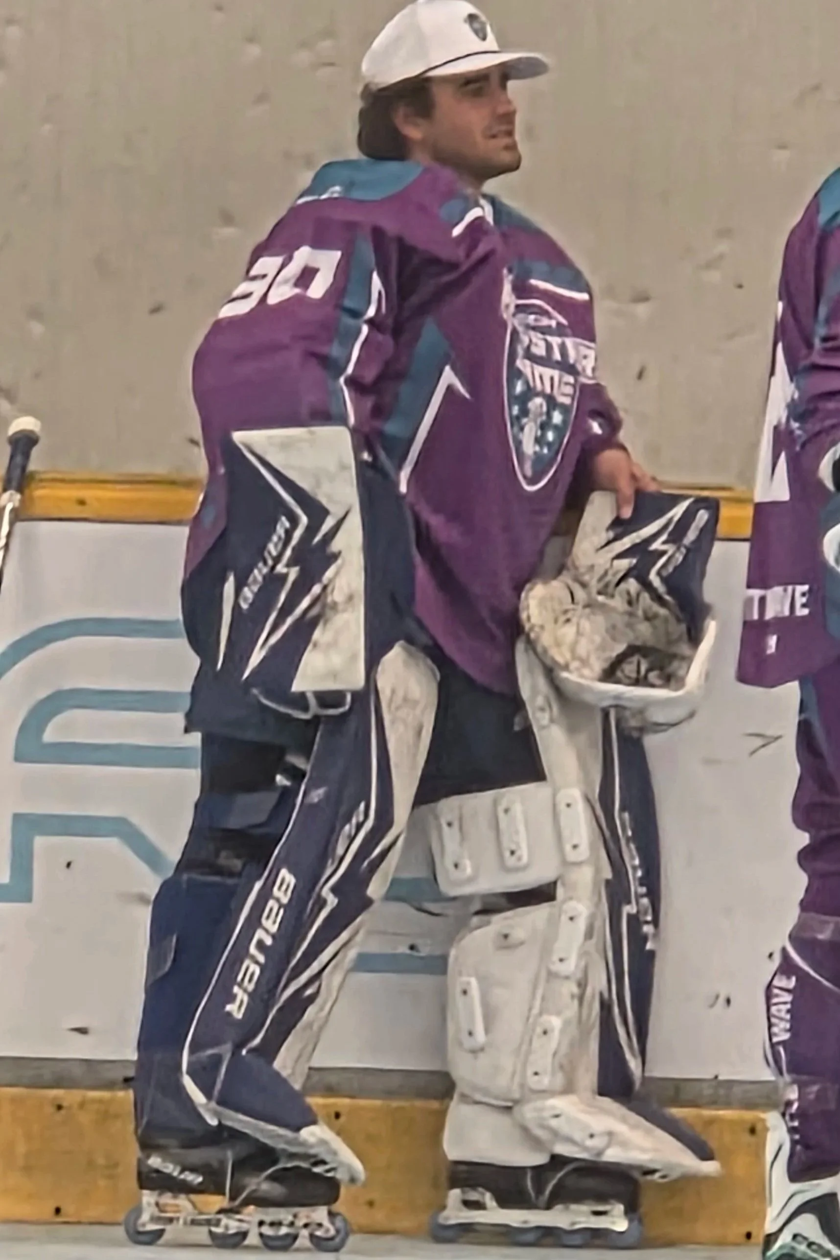 Hockey player wearing a purple jersey and goalie equipment, standing inside an ice rink with a wooden barrier behind him.