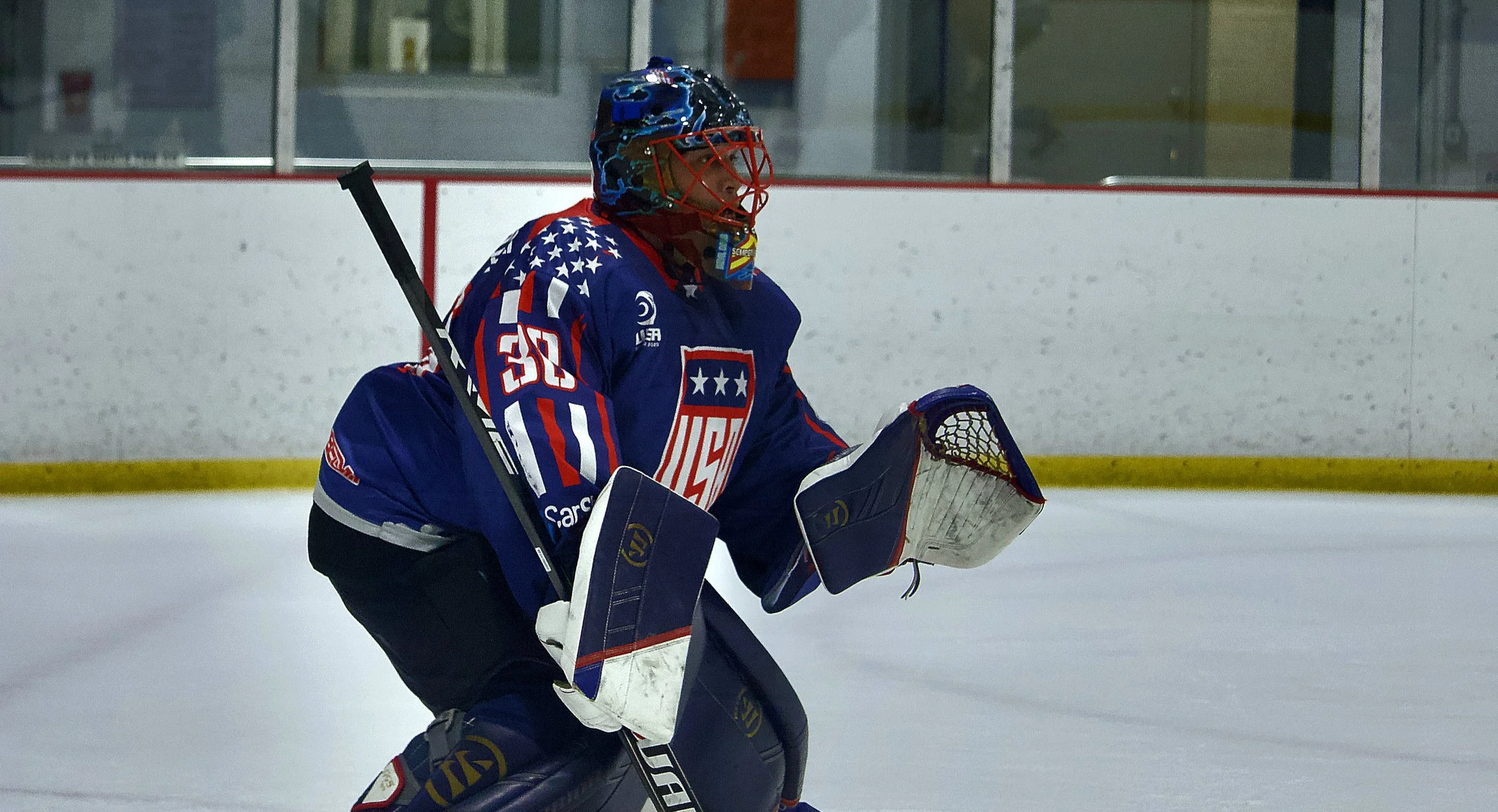 A hockey goaltender in a blue jersey with red, white, and blue patriotic stars and stripes design, wearing a blue helmet with a colorful design, pads, and gloves, is on the ice in front of the goal during a game.