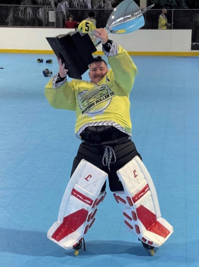 A man wearing goalie's equipment and a yellow hockey jersey is holding a trophy above his head on an indoor roller hockey rink, celebrating a victory.