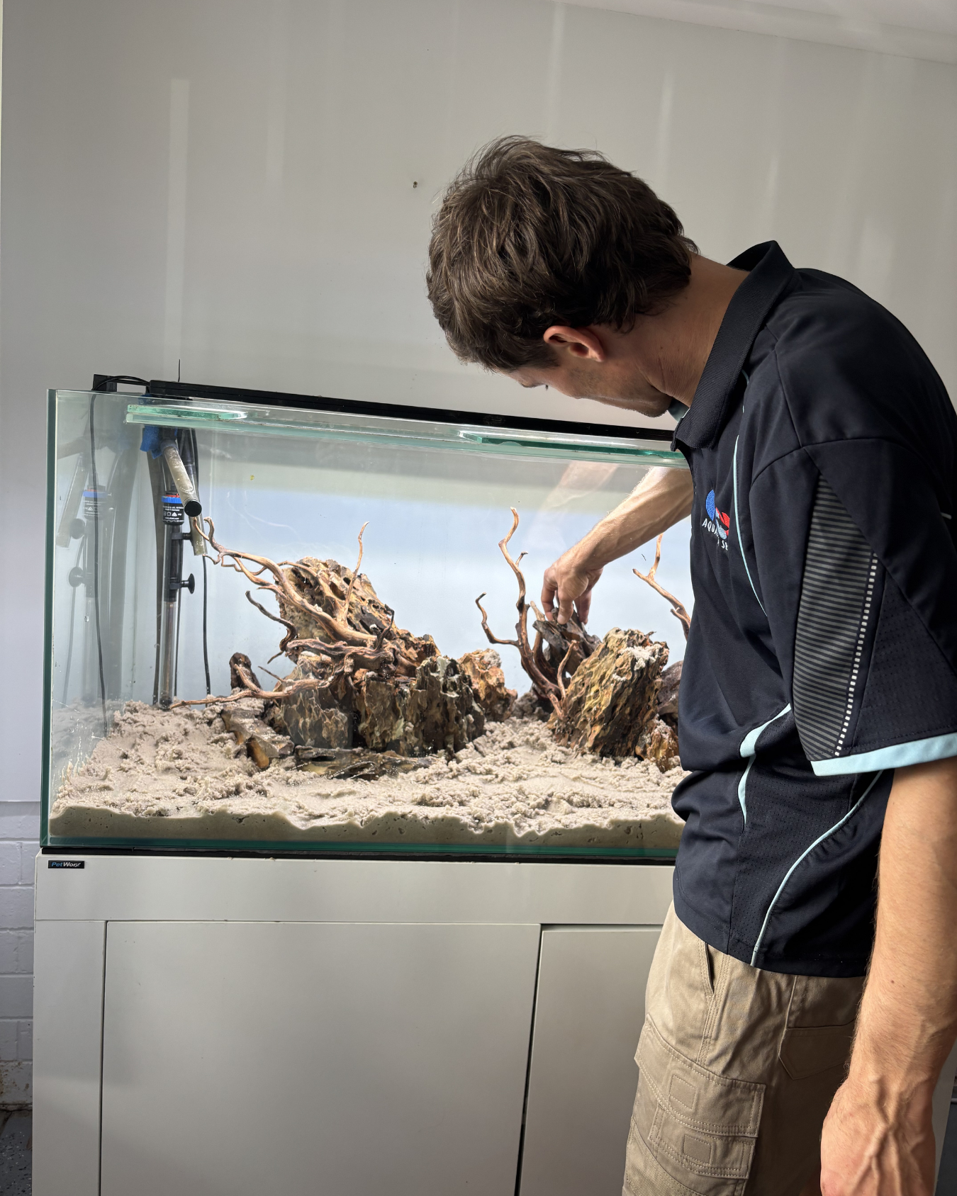 A man in a black shirt and tan shorts is adjusting a desert terrarium with sand, rocks, and dry, twisted branches inside a glass tank.