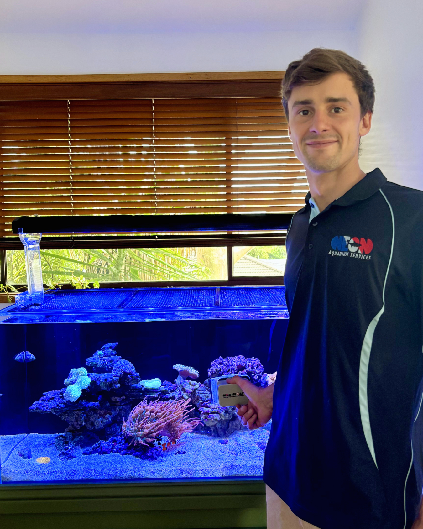 A man in a black polo shirt with the logo 'AQUARIUM SERVICES' stands next to a large saltwater aquarium filled with corals and marine life. The background features window blinds with sunlight filtering through.