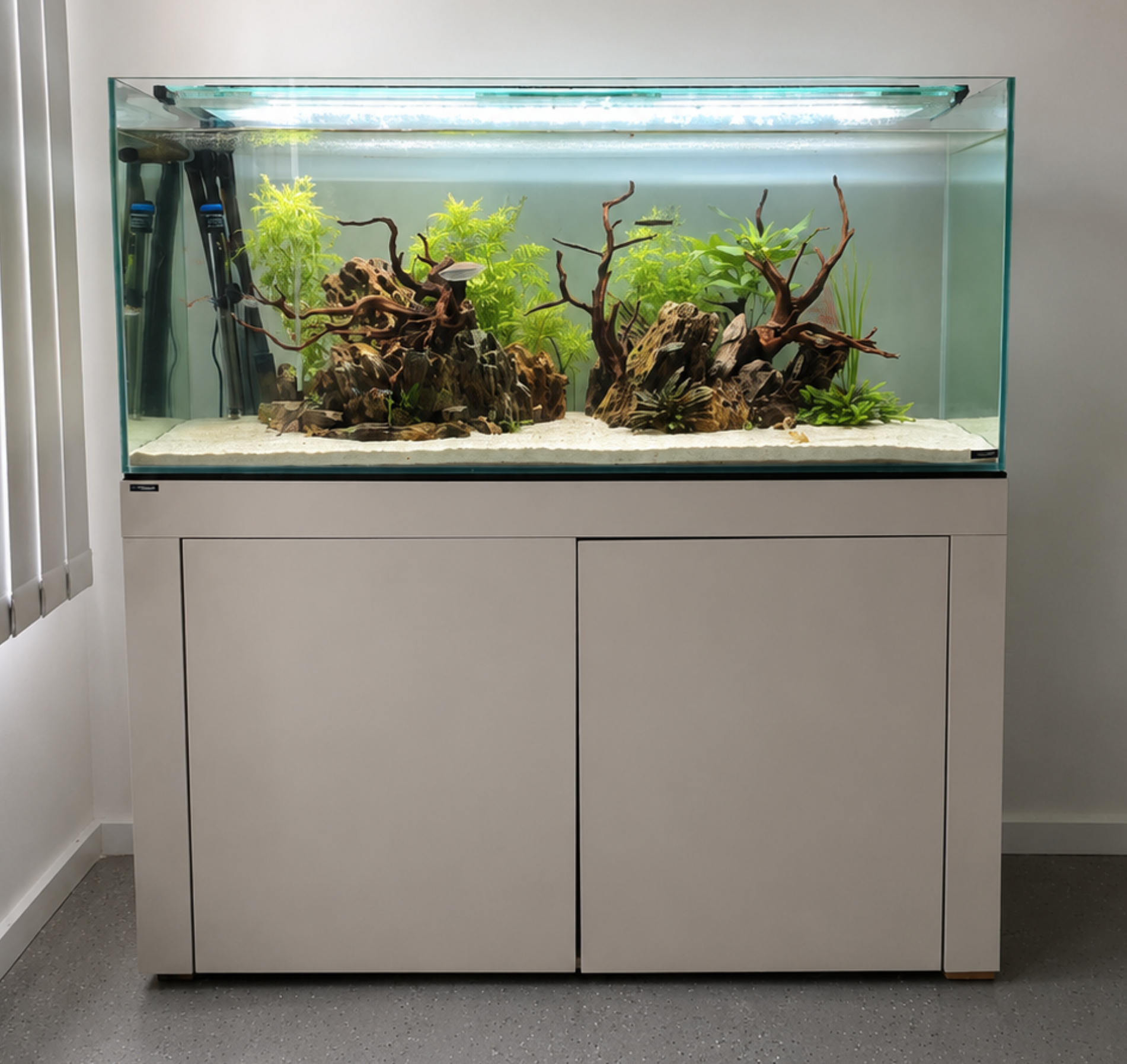 A glass aquarium with aquatic plants and driftwood decorations, placed on a white cabinet in a room with gray speckled flooring and white walls. The tank has a lighting system on top.