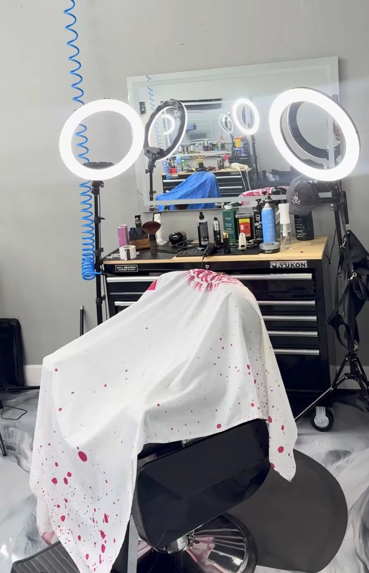 Barber's station with ring lights, mirror, and barbering tools, covered with a cloth with pink paint splatters, and a barber chair in front.