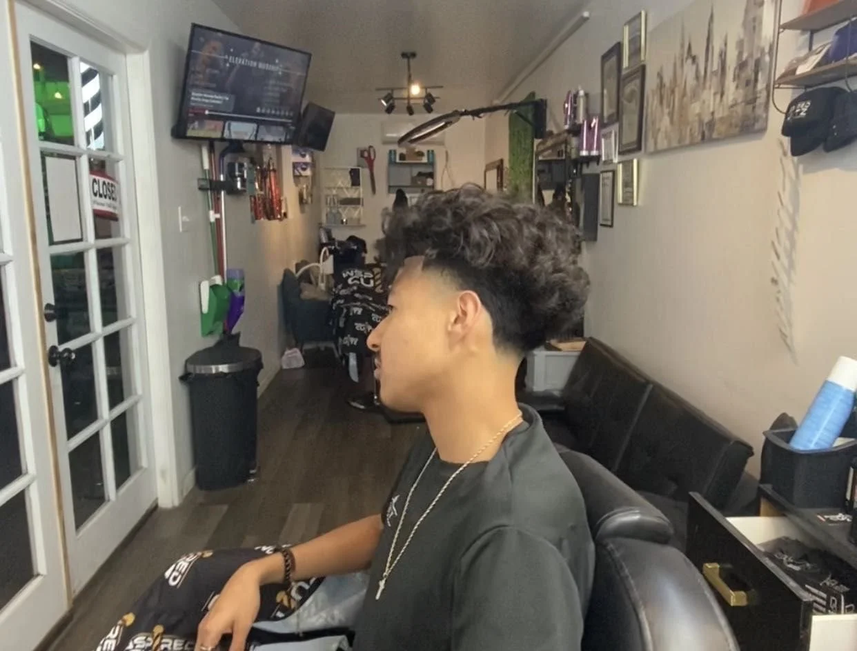 Interior of a barber shop with a young man sitting on a black chair, having a fresh haircut. The wall behind features multiple framed photos and artwork, and a flat-screen TV is mounted high on the wall. There is a door with glass panels to the left.