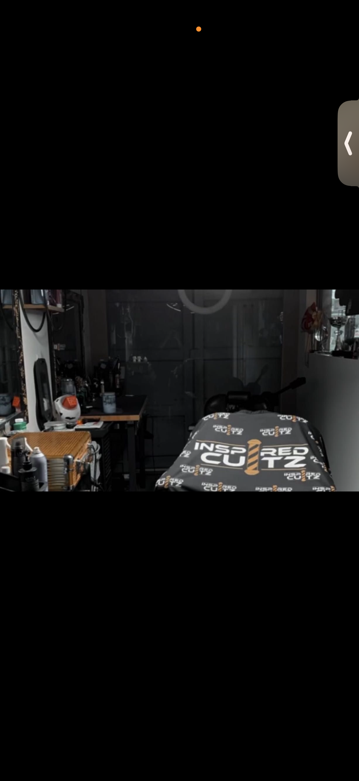 A room with a bed covered in a black and white blanket that has the logo 'Insured Cutz'. The room has various items on the shelves, a work table with equipment, and a garage-style door in the background.