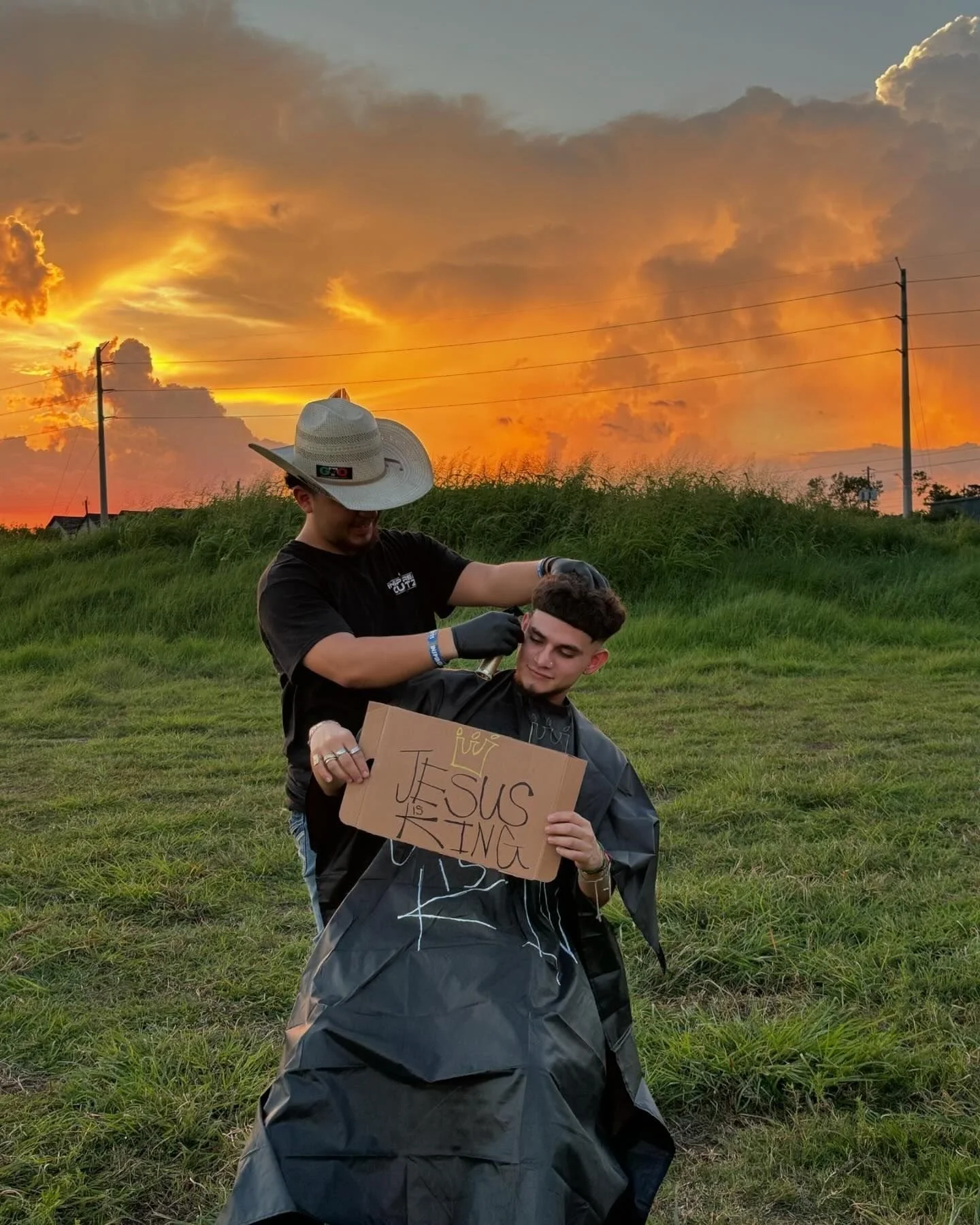 A person receiving a haircut outdoors during sunset, with a person holding a sign that reads 'Jesus is King' while sitting on the grass.