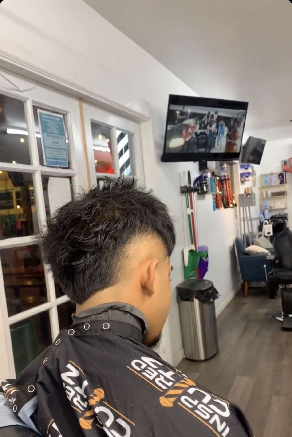A person with a short, styled designed haircut sitting in a barber shop with televisions on the wall and various hair care products.