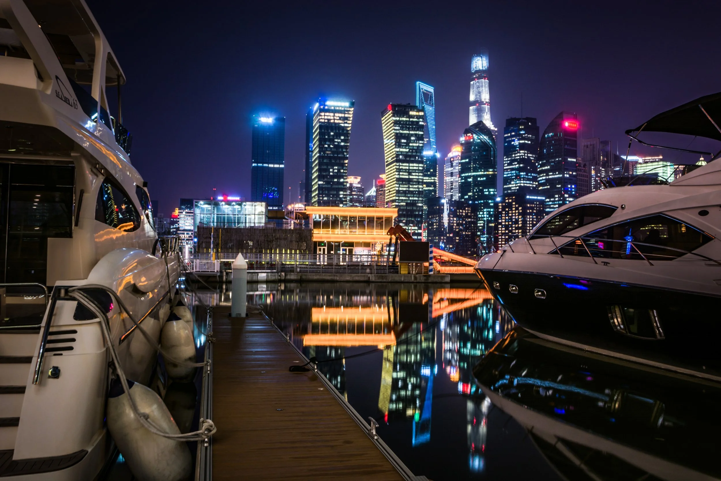 Nighttime city skyline with tall illuminated skyscrapers and reflections on the water. Foreground shows docked yachts and boats at a marina.