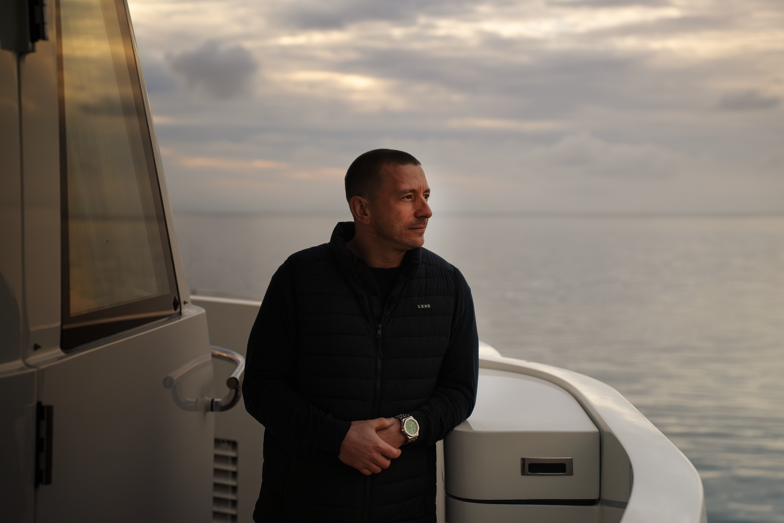 A man wearing a black quilted jacket and a watch stands on a boat, looking out at a cloudy sky over the ocean. Part of the boat's structure is visible on the left and front side of the image.