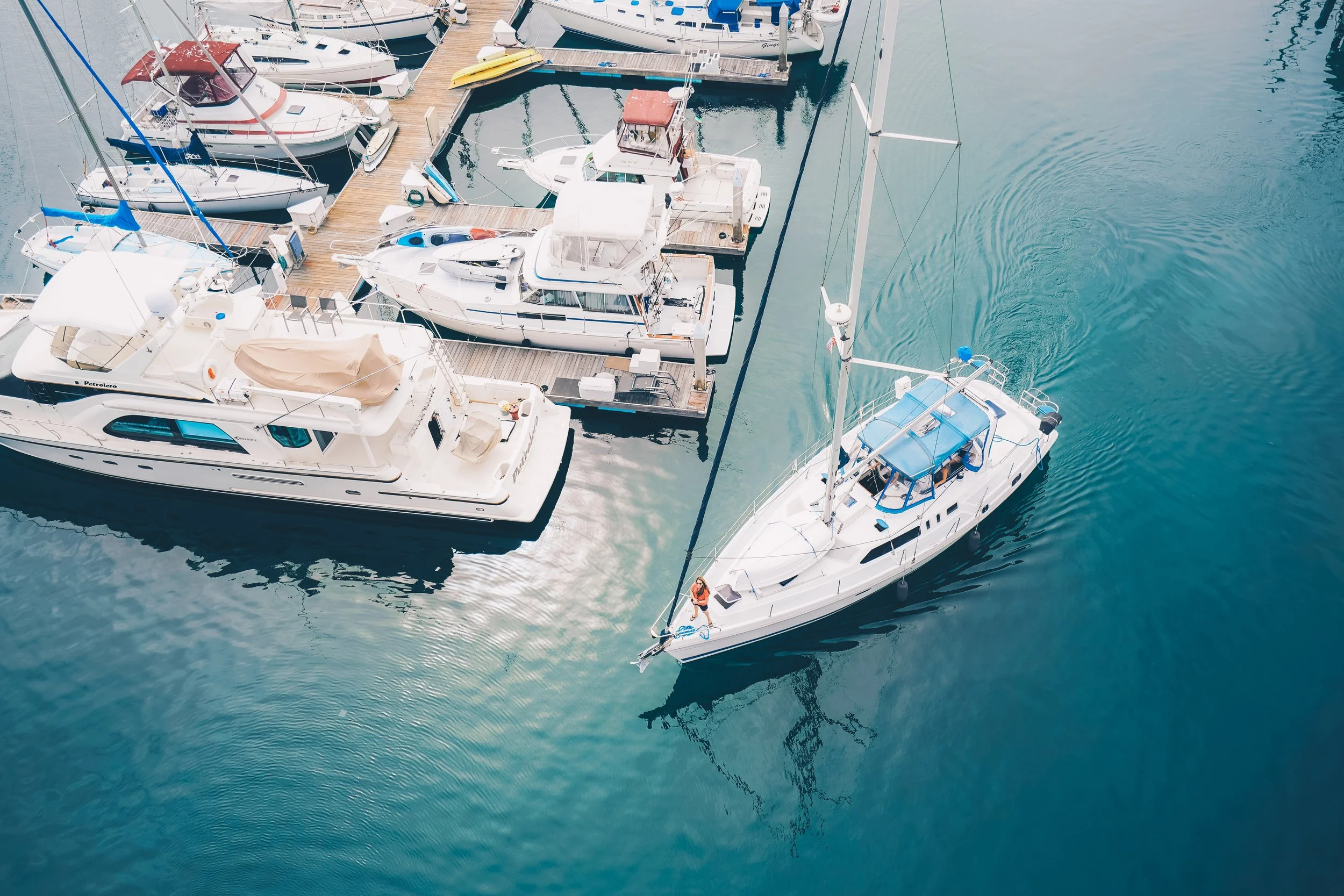 An aerial view of a marina with several sailboats and motorboats docked along floating docks. The water is calm, creating a mirror-like reflection of the boats.