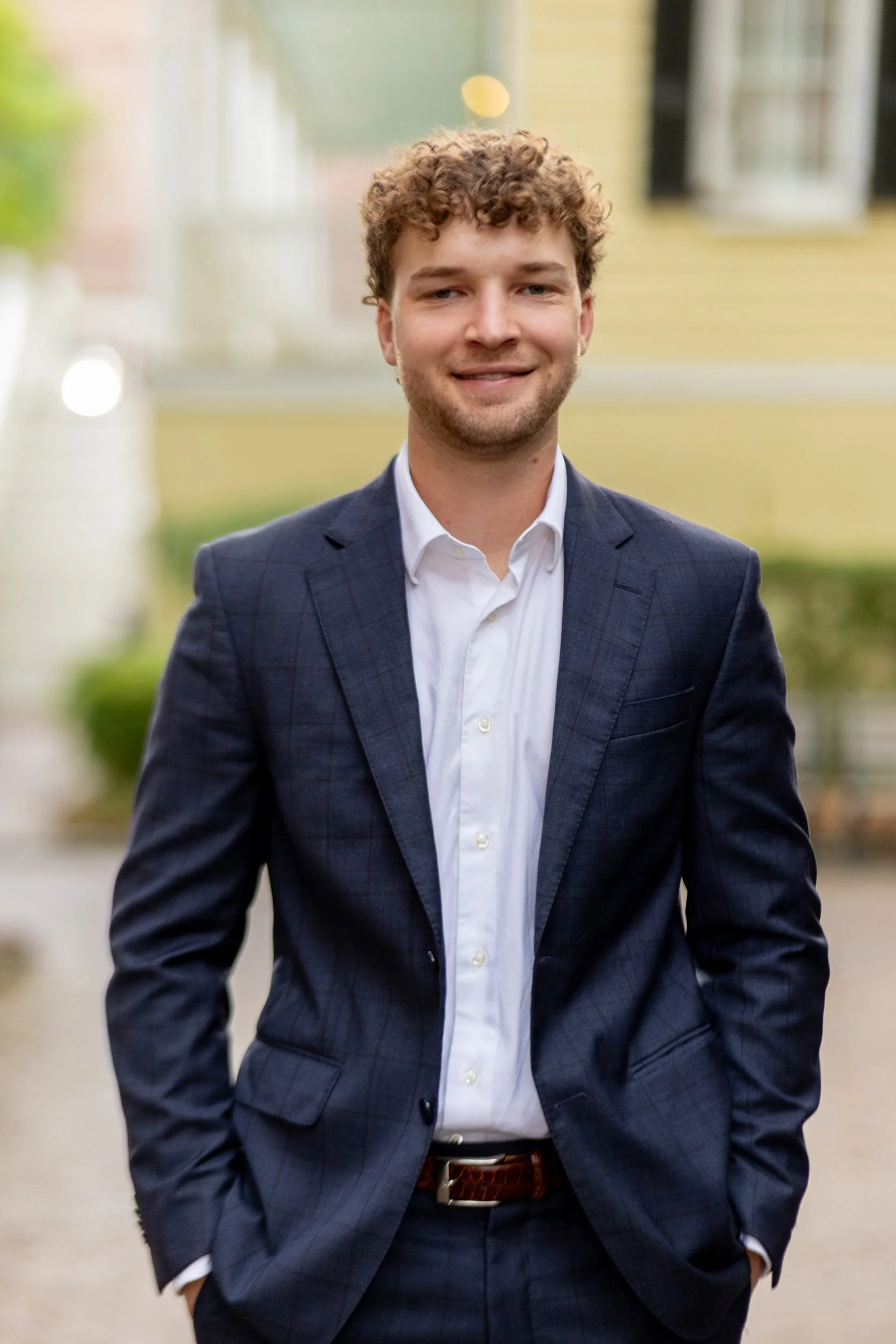A young man with curly brown hair wearing a dark suit jacket over a white dress shirt, standing outdoors with blurred background of a yellow house and greenery.