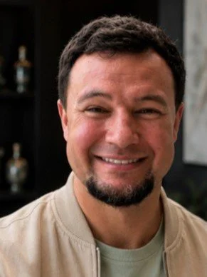A smiling man with dark hair and a goatee, wearing a beige jacket and a light-colored shirt, standing indoors.
