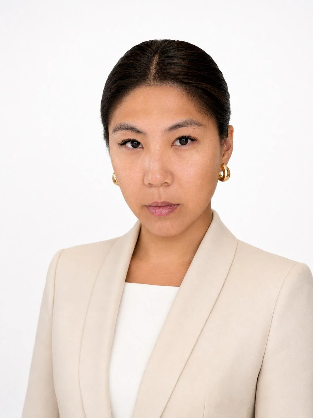A woman with dark hair styled back, wearing a cream-colored blazer, white top, and gold hoop earrings, staring confidently into the camera against a plain white background.