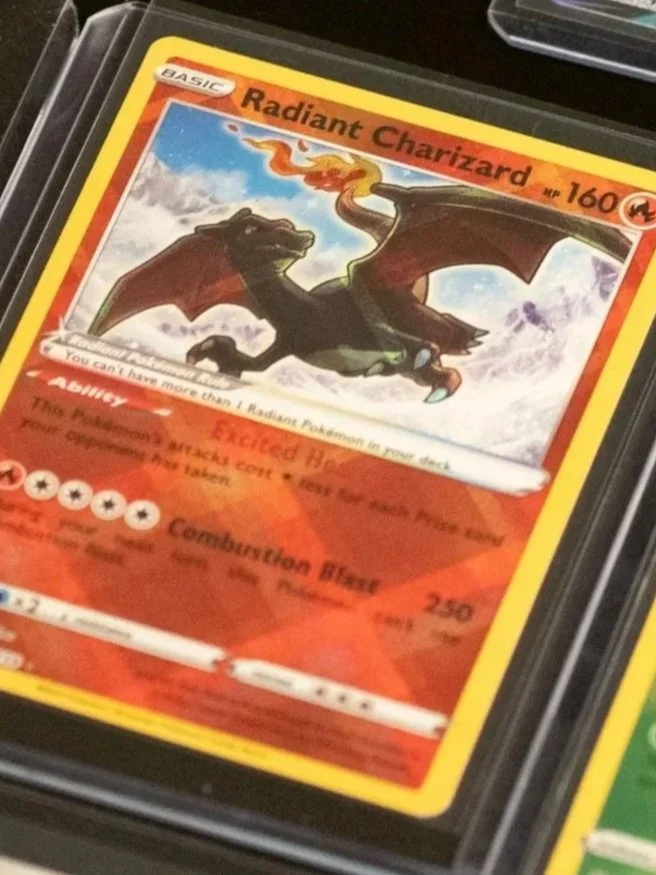 A Pokémon card labeled Radiant Charizard with 160 HP, depicting Charizard breathing fire. The card is part of the Basic Pokémon series and features an ability called 'Speed Heat' and an attack called 'Combustion Blast' with 250 damage. The card has a fiery orange and red background.