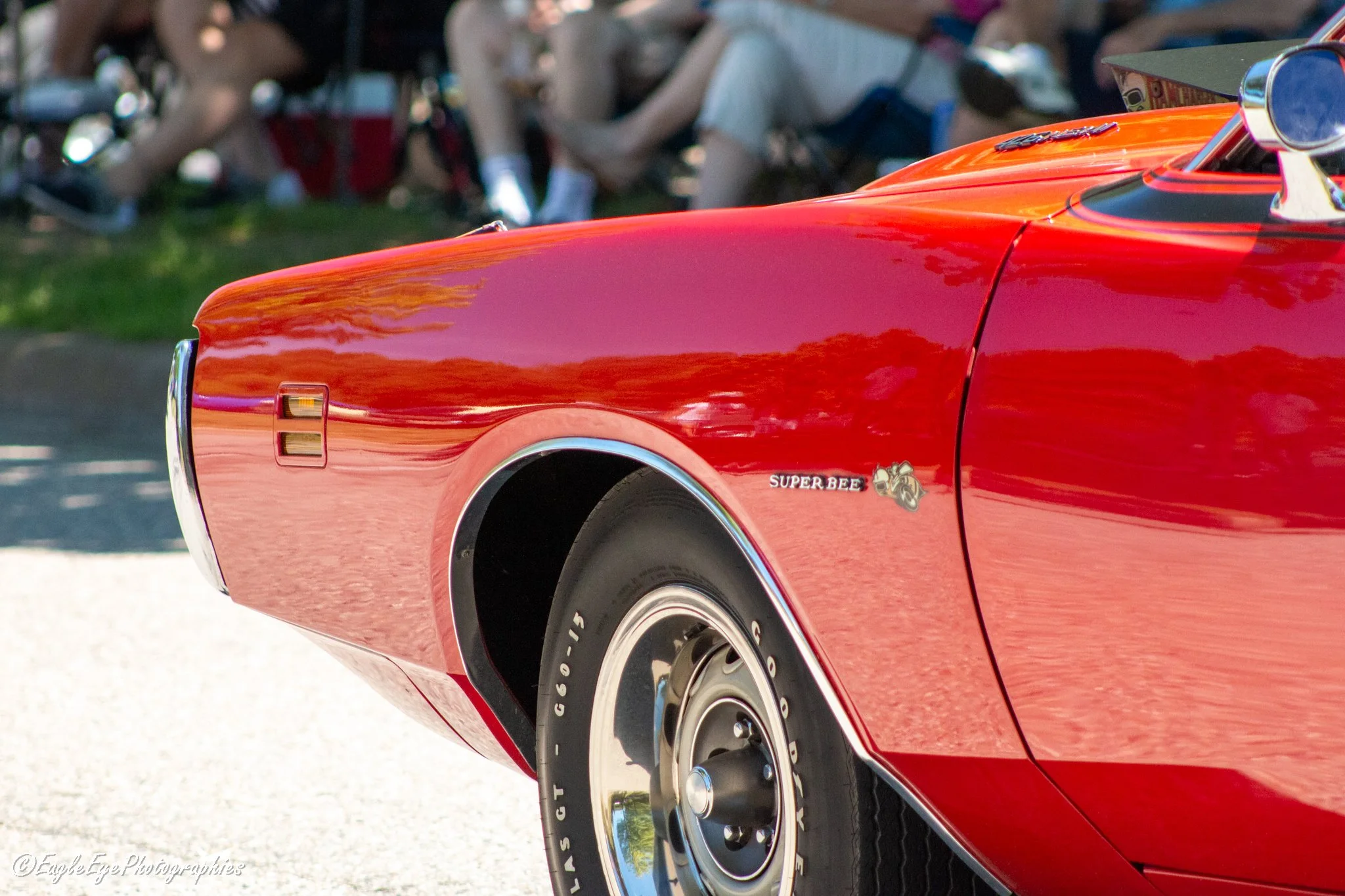 Close-up of a red classic muscle car, showing the front left side with the wheel, fender, and part of the hood. The car has a shiny finish with a Super Bee emblem on the side.