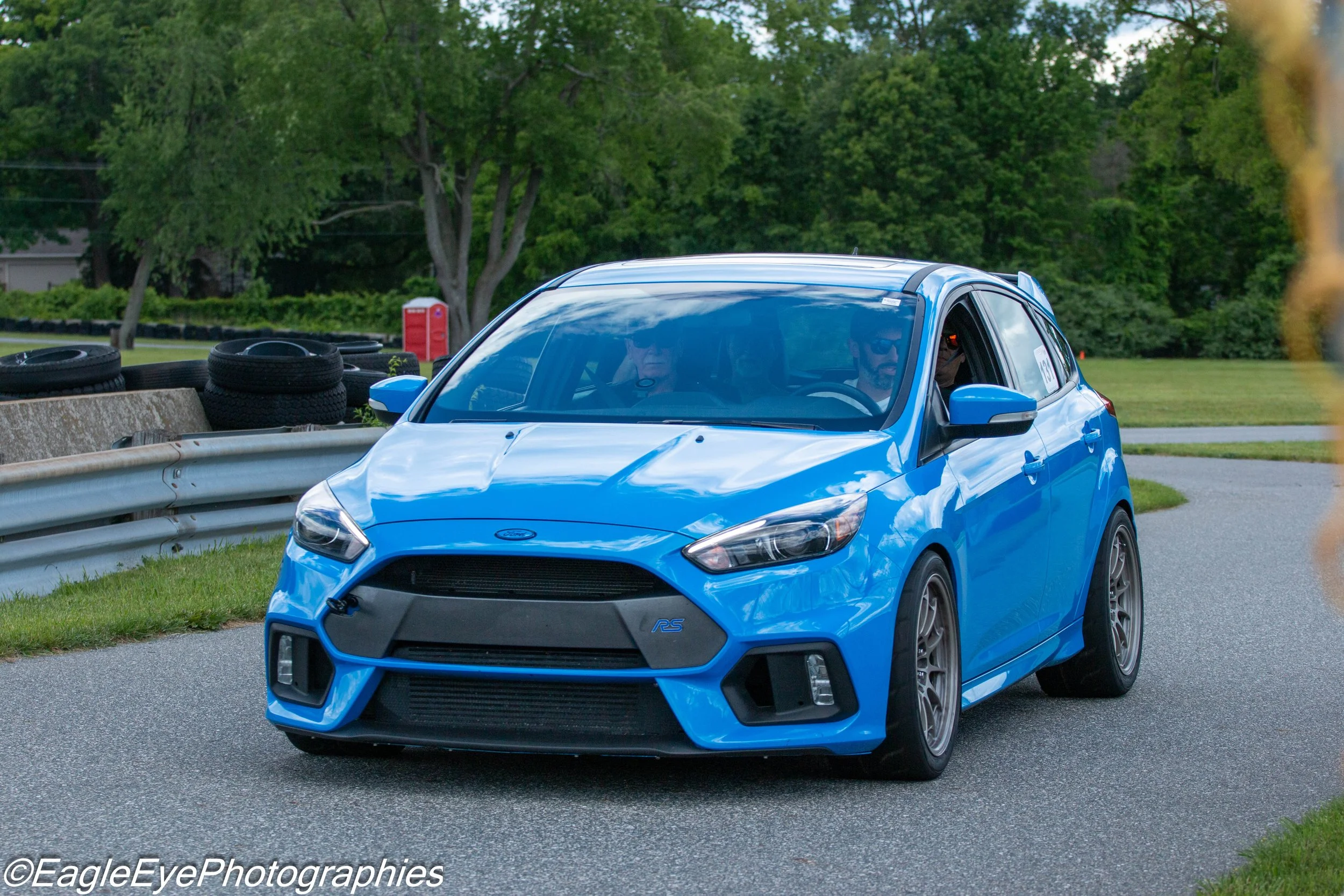 A blue Ford car driving on a paved road with a green wooded background and tire barriers on the side. Two people are inside the car, both wearing sunglasses.