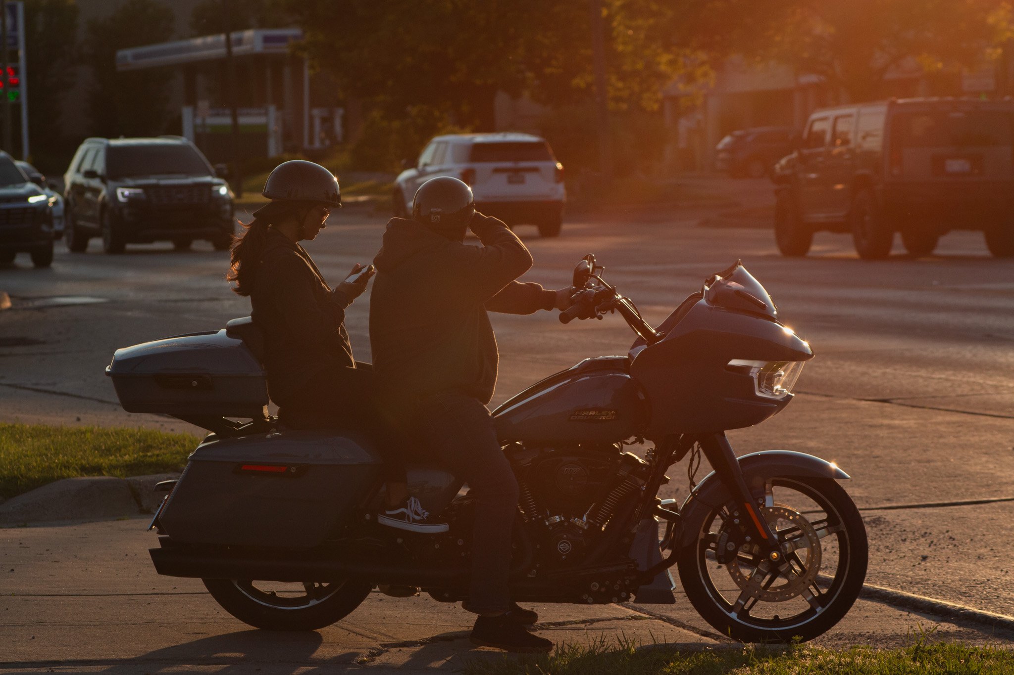 Two people wearing helmets are sitting on a motorcycle at sunset on a city street, with cars and trees in the background.