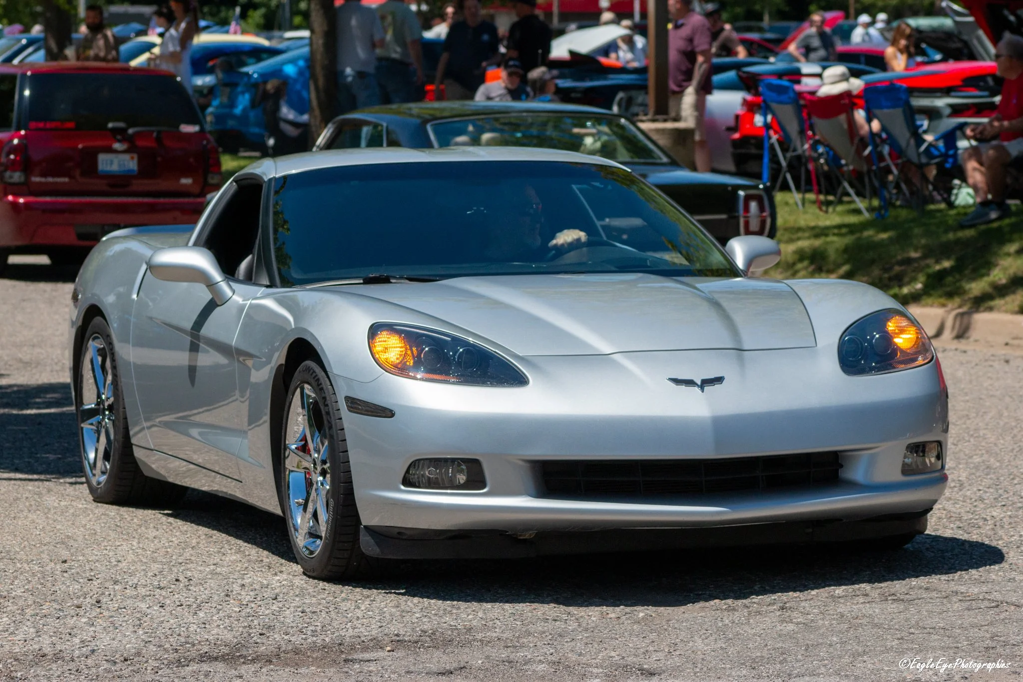 A silver Chevrolet car parked at a car show with other vehicles and people in the background.