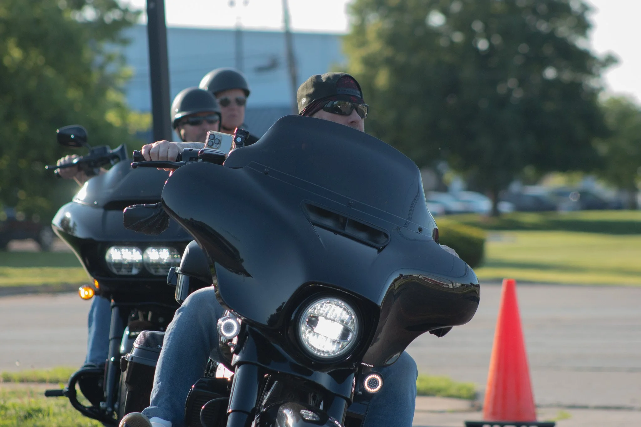 Three motorcyclists wearing helmets and sunglasses riding in a line outdoors near orange traffic cones, with trees and a blue sky in the background.