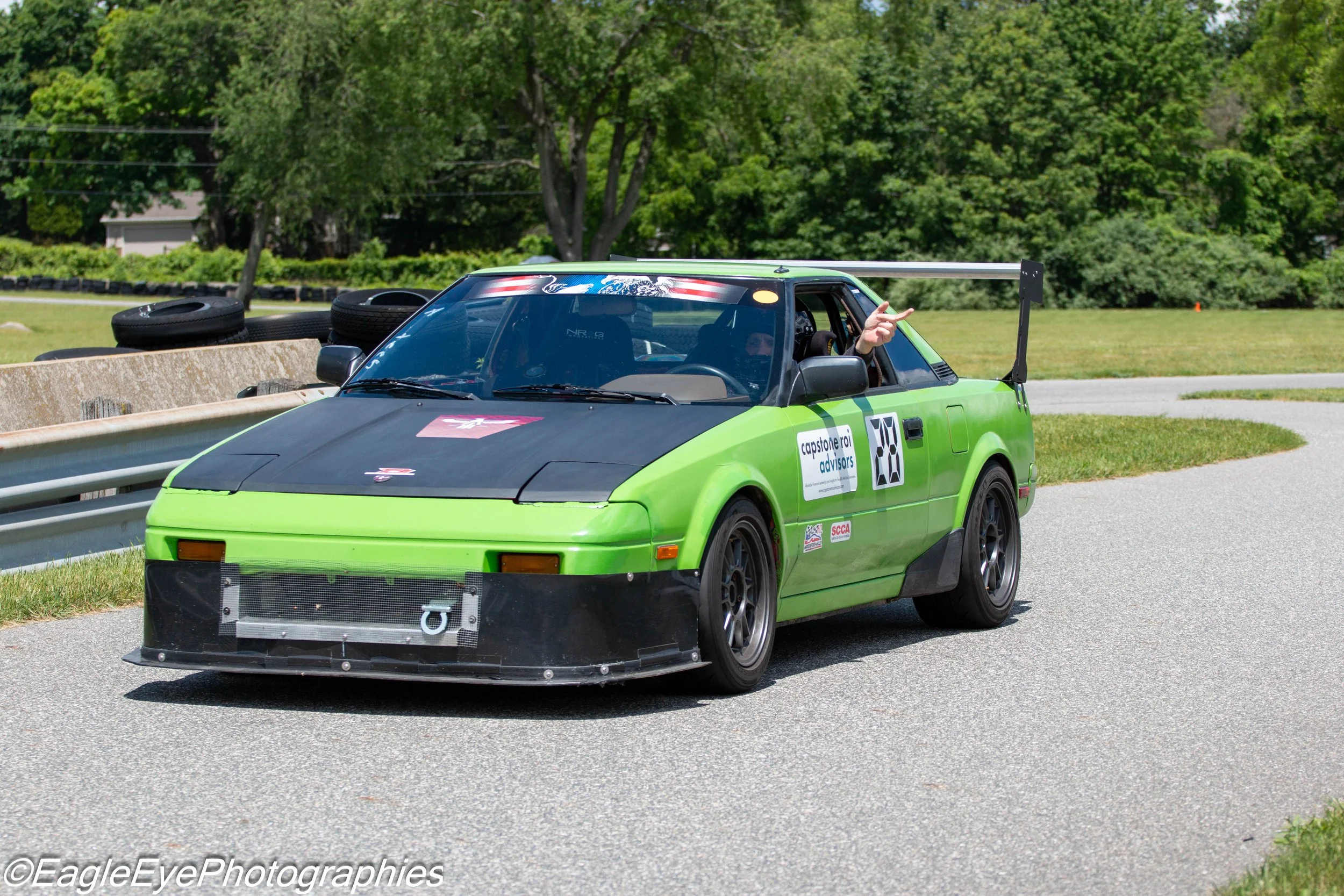 A green race car on a track with a person inside pointing with their finger. The car has a large rear wing, tires on the back, and a front grille.